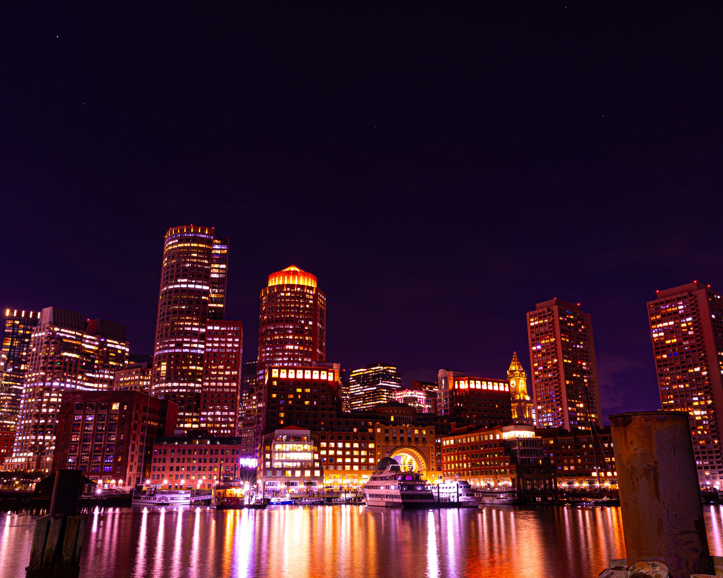 Nighttime city skyline with illuminated skyscrapers reflecting on water, including a tall round building, a clock tower, and boats docked along the water.