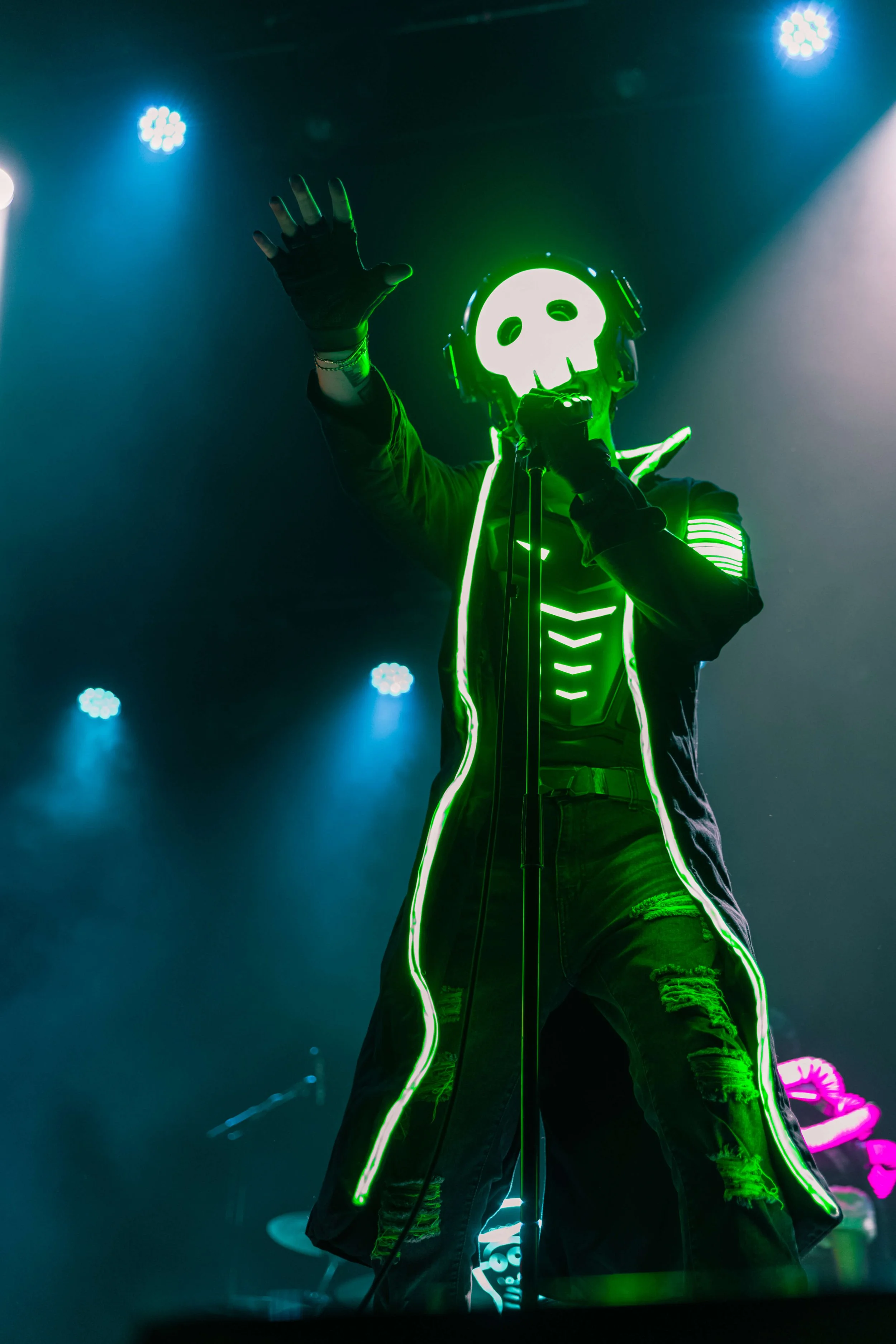Performer wearing black jacket with glowing green neon skeleton design and skull mask on stage, raising hand in a dark setting with stage lights.