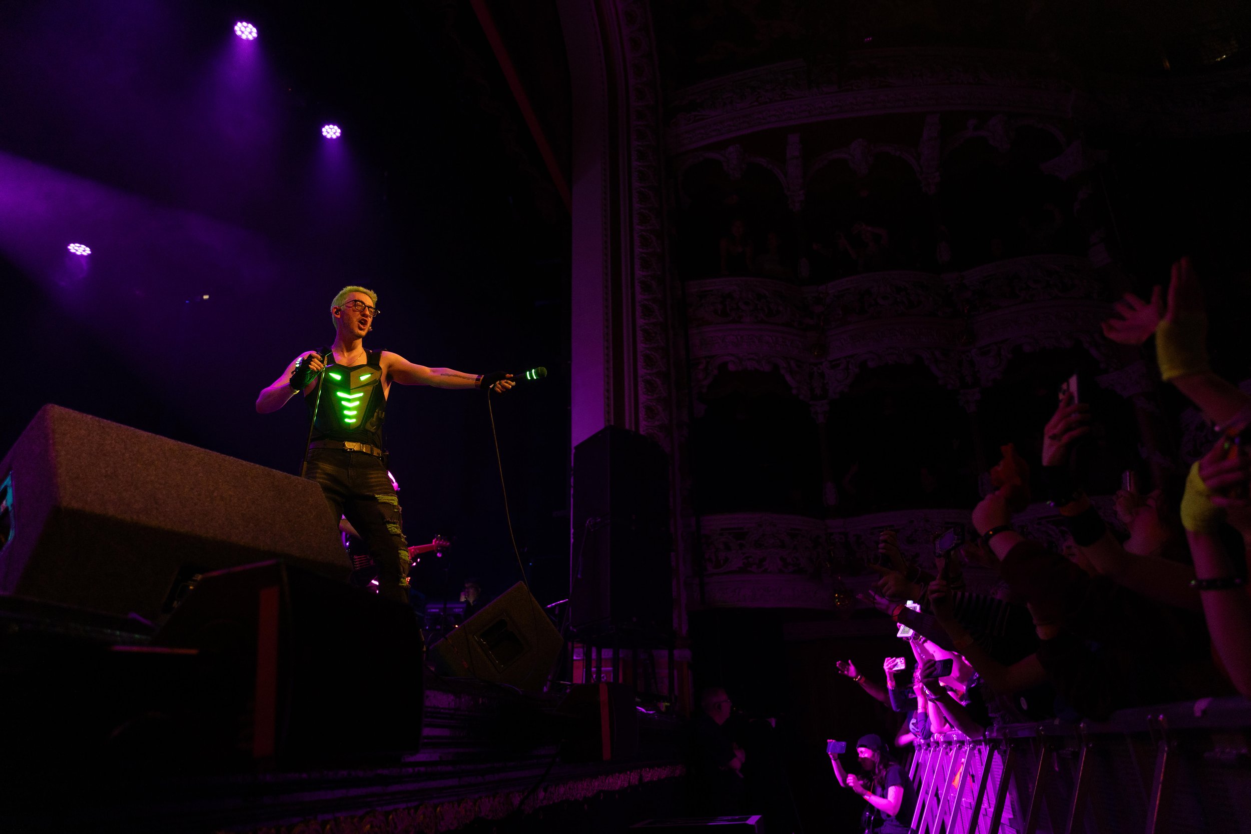 Performer on stage with light-up costume holding a microphone, audience members with light-up accessories reaching towards the stage, ornate theater interior with balconies and decorative moldings, purple stage lighting