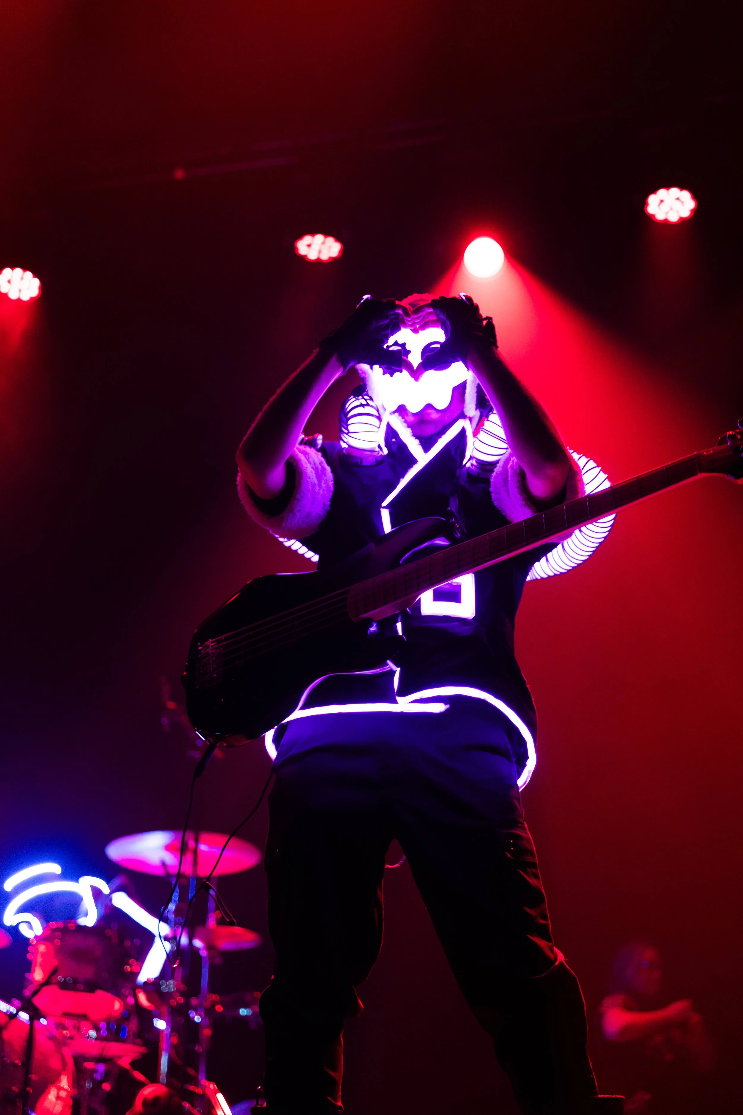 A performer wearing an LED costume with glowing rings and a mask, playing a guitar on stage with red lights and a neon sign in the background.