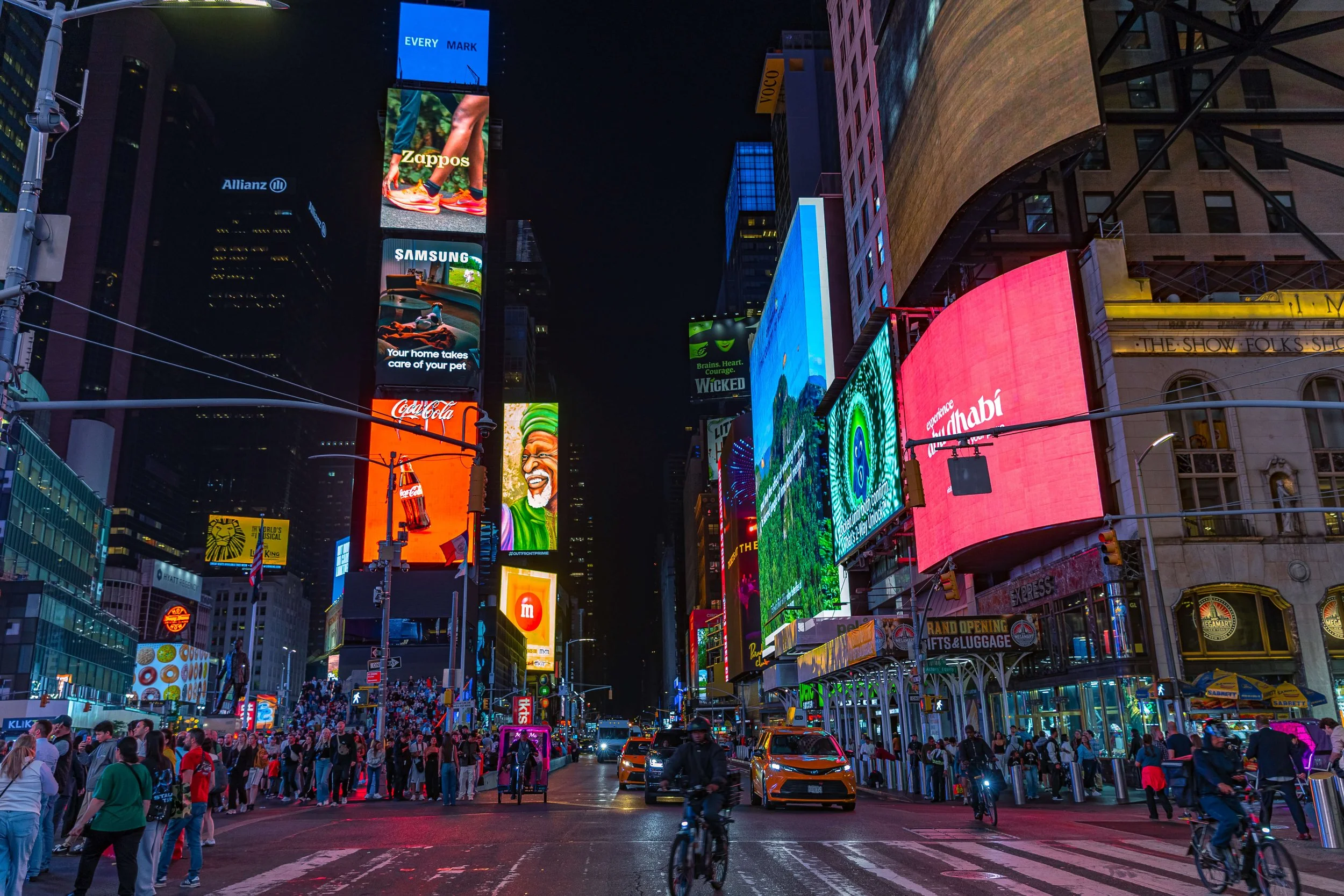 Nighttime scene in Times Square, New York City with bright digital billboards, a crowd of pedestrians, yellow taxis, cyclists, and surrounding buildings.