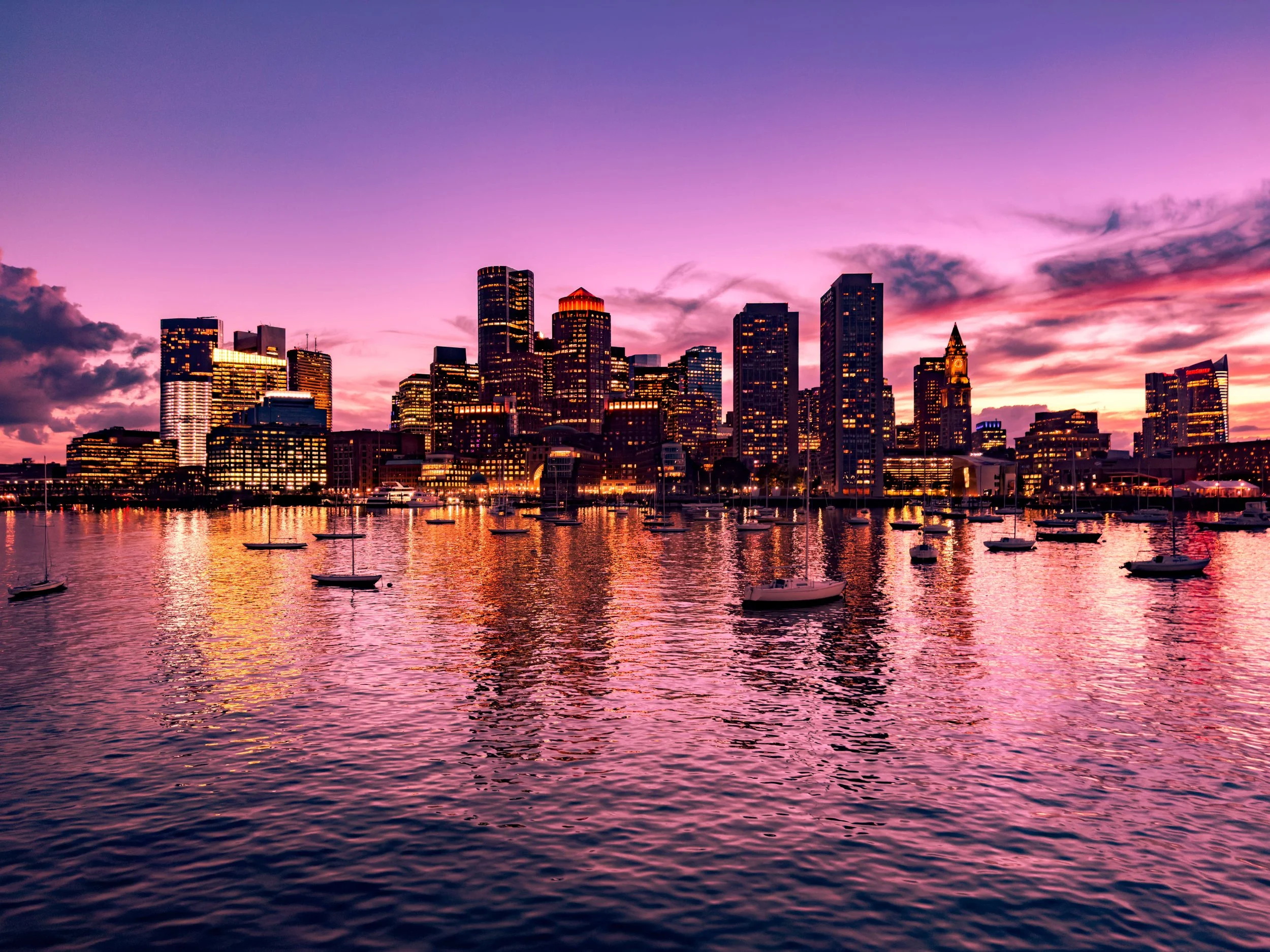 City skyline at sunset with tall buildings reflecting in a harbor filled with boats.