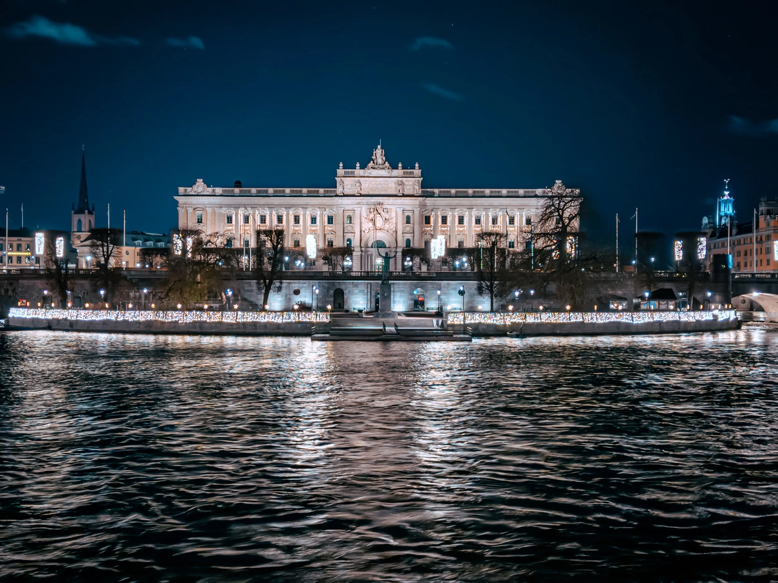 A historic building illuminated at night, situated along a river with reflections on the water.