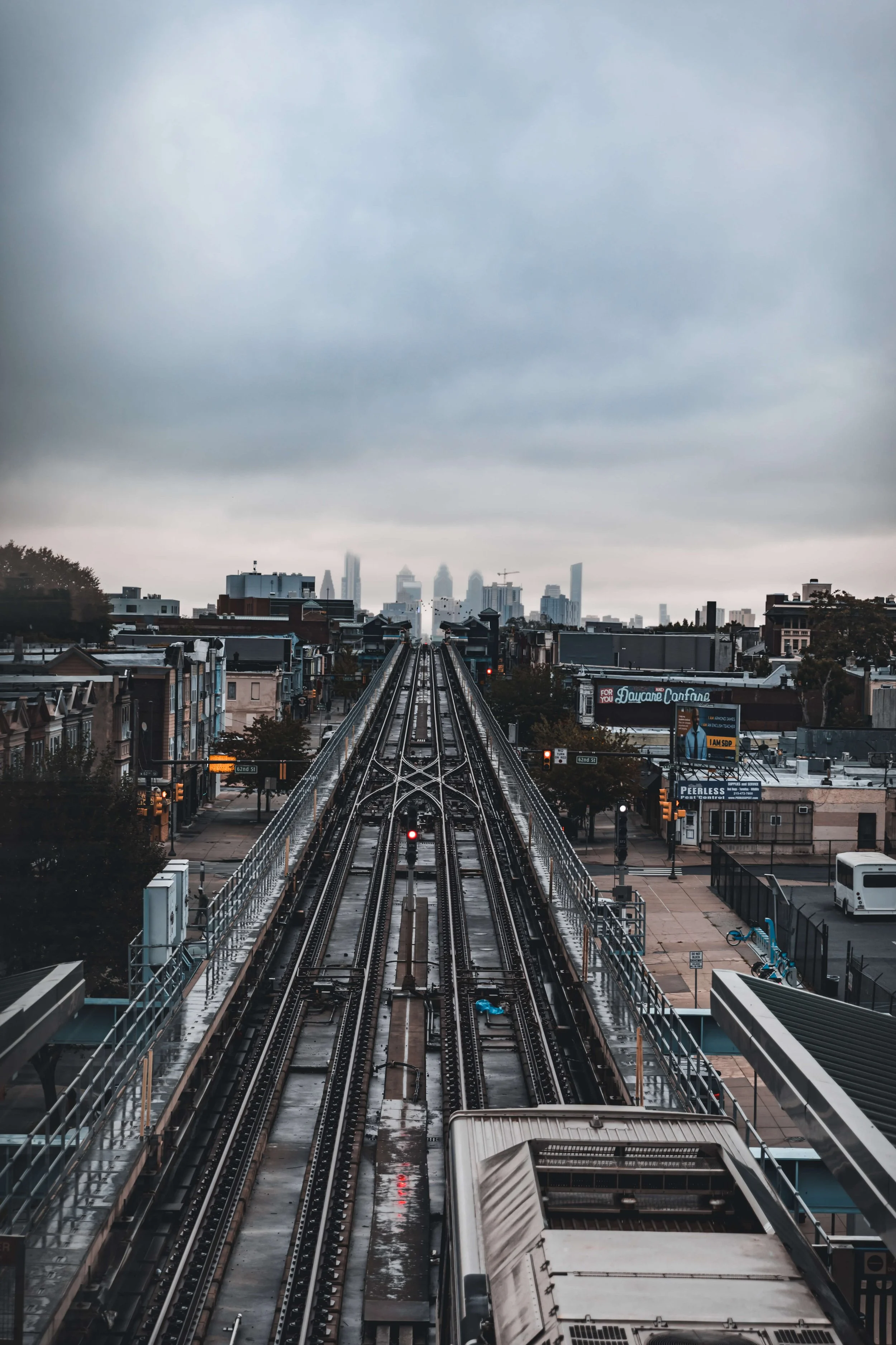 View of an elevated train track leading into a city skyline on a dreary, overcast day, with rain-soaked streets below.