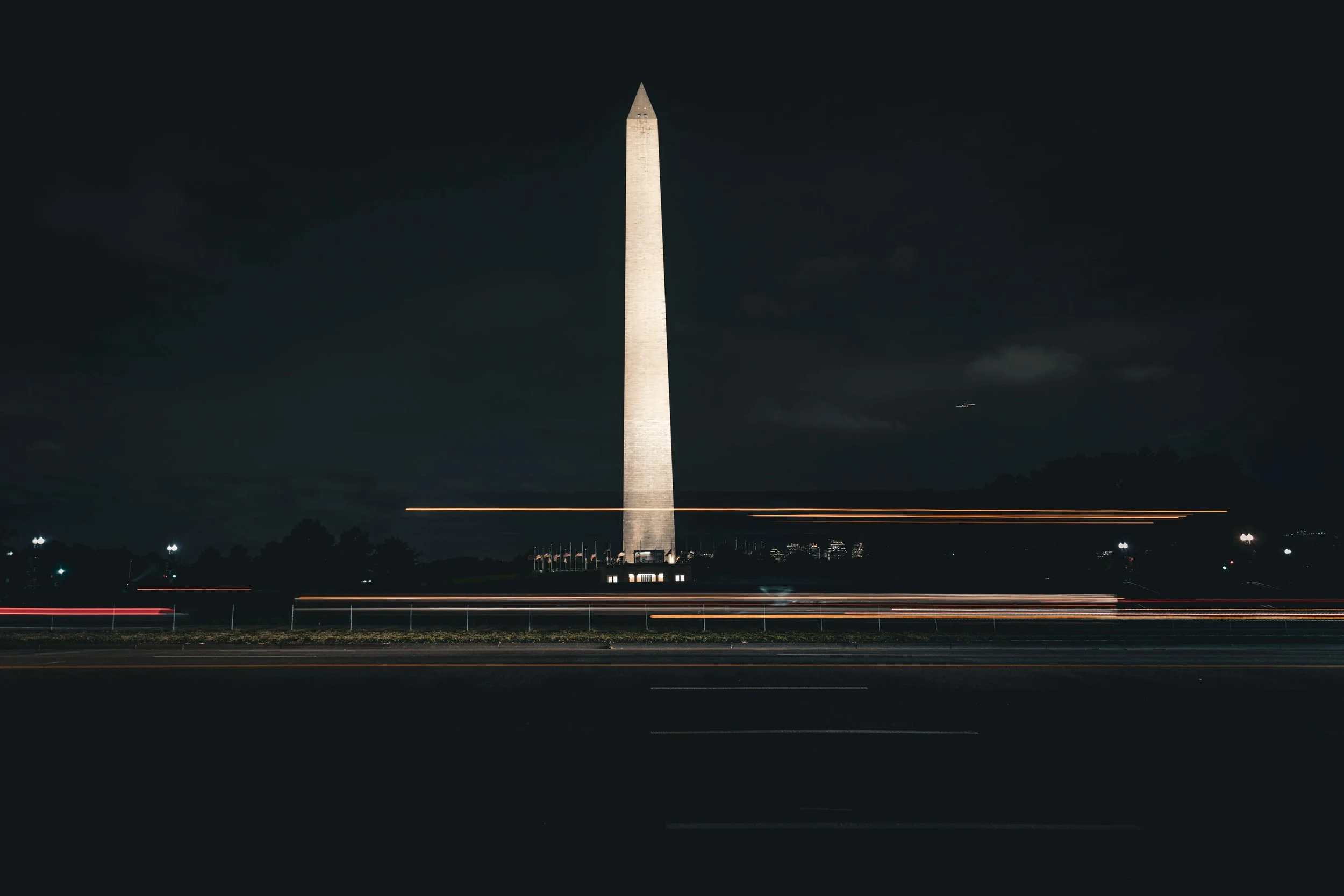 Nighttime view of the Washington Monument illuminated with light, with light trails from passing vehicles in the foreground.