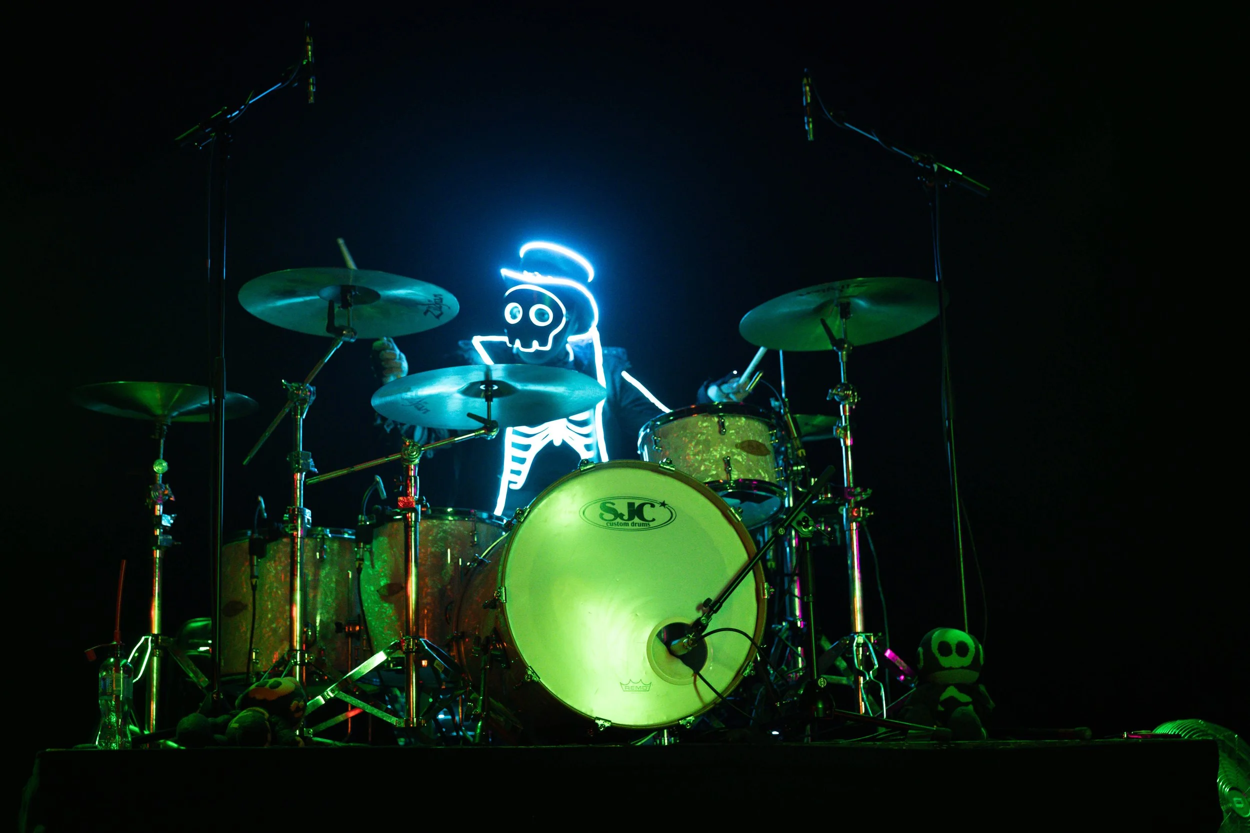 A drum set with drumsticks, adorned with skeleton and skull decorations, and a figure with a neon glowing skeleton face behind the drums, with neon skeleton outlines glowing on the figure.