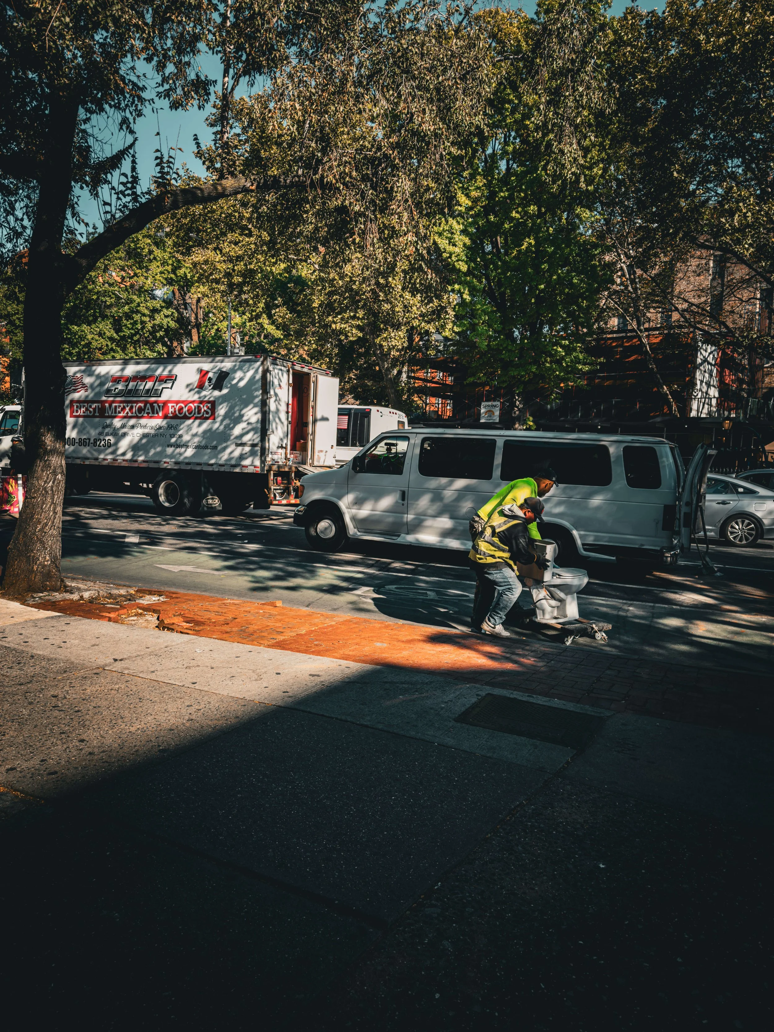 Two workers in fluorescent safety vests working on the sidewalk near a white van and a moving truck on a city street, with trees overhead and parked cars in the background.