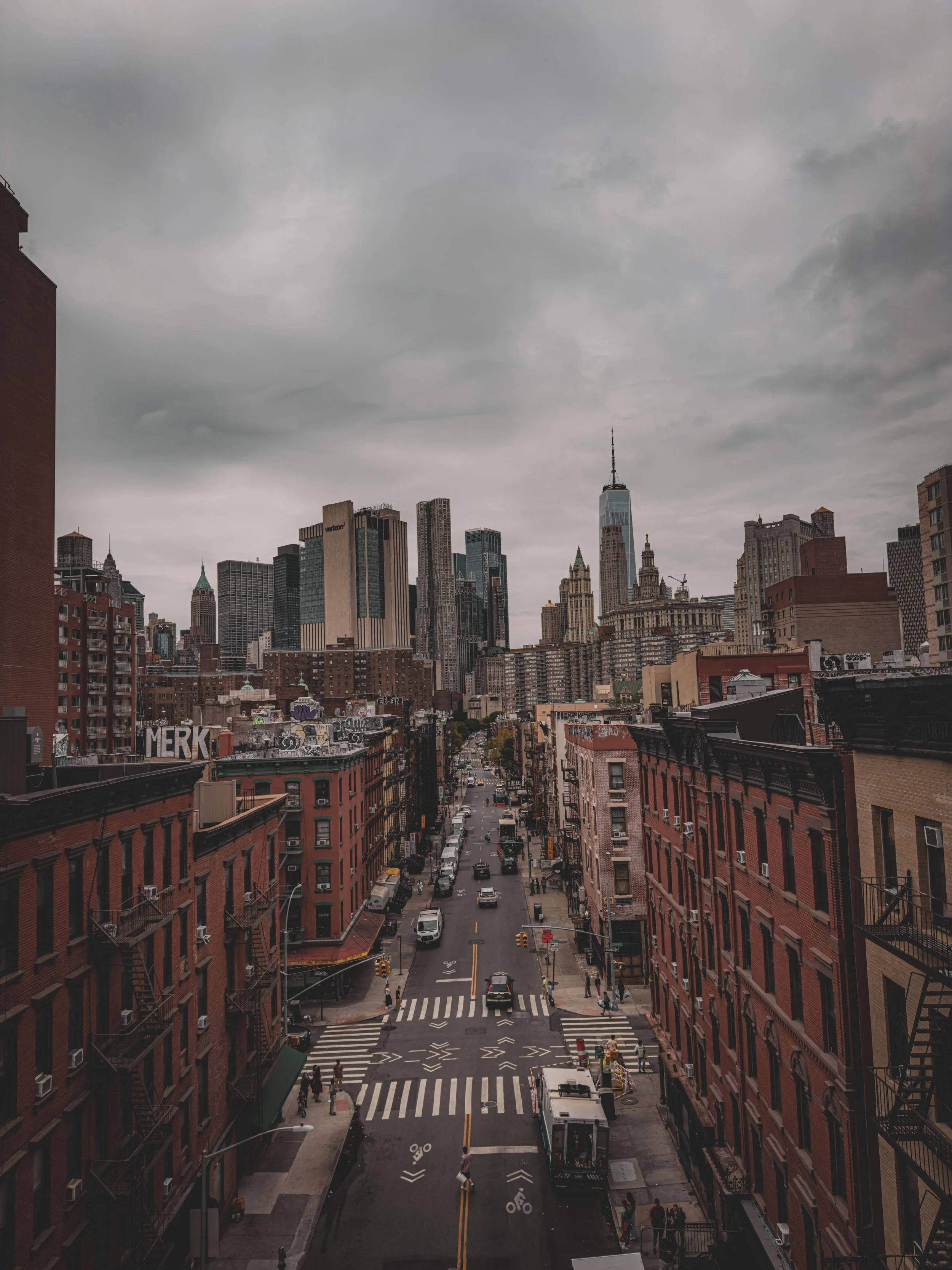City street with crosswalks, cars, and pedestrians in front of red brick buildings, with a skyline of tall skyscrapers, including One World Trade Center, under a cloudy sky
