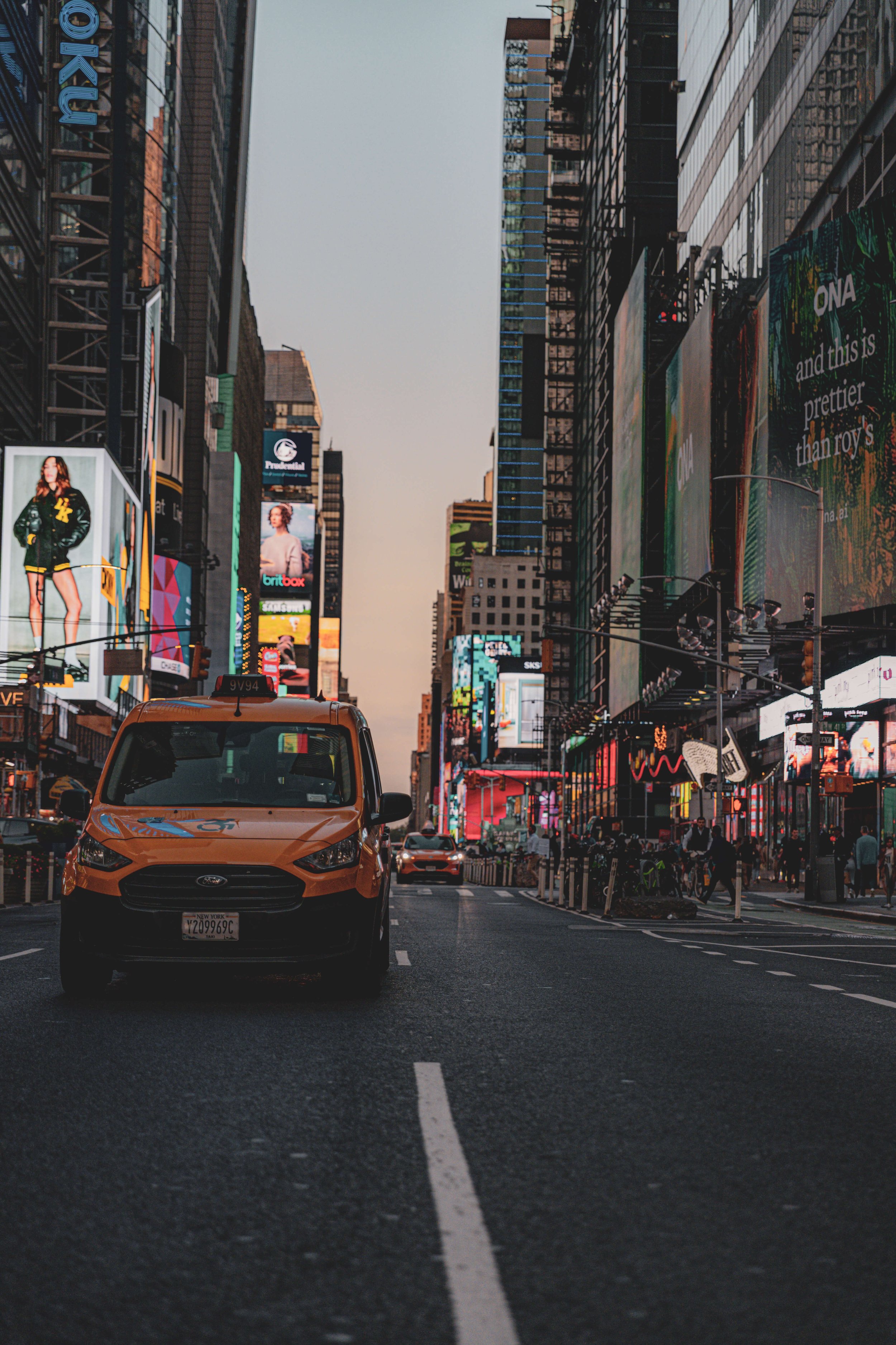 A yellow taxi cab driving on a busy city street with tall buildings and large digital billboards at dusk in Times Square, New York City.