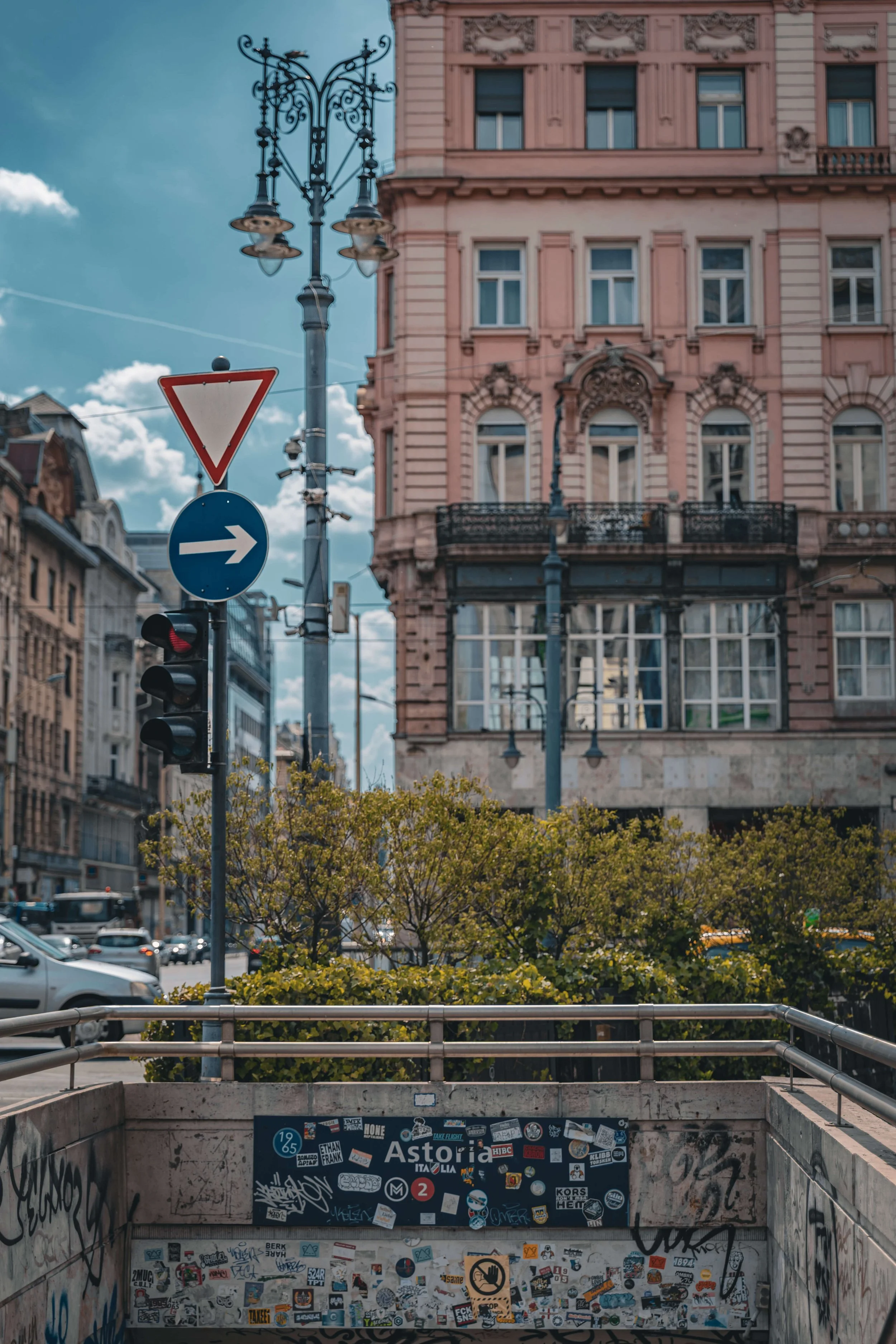 Street scene with a pink ornate building, traffic lights, street signs, and a patterned blue sky with clouds. The foreground features a graffiti-covered entrance to an underground passage with numerous stickers and posters.