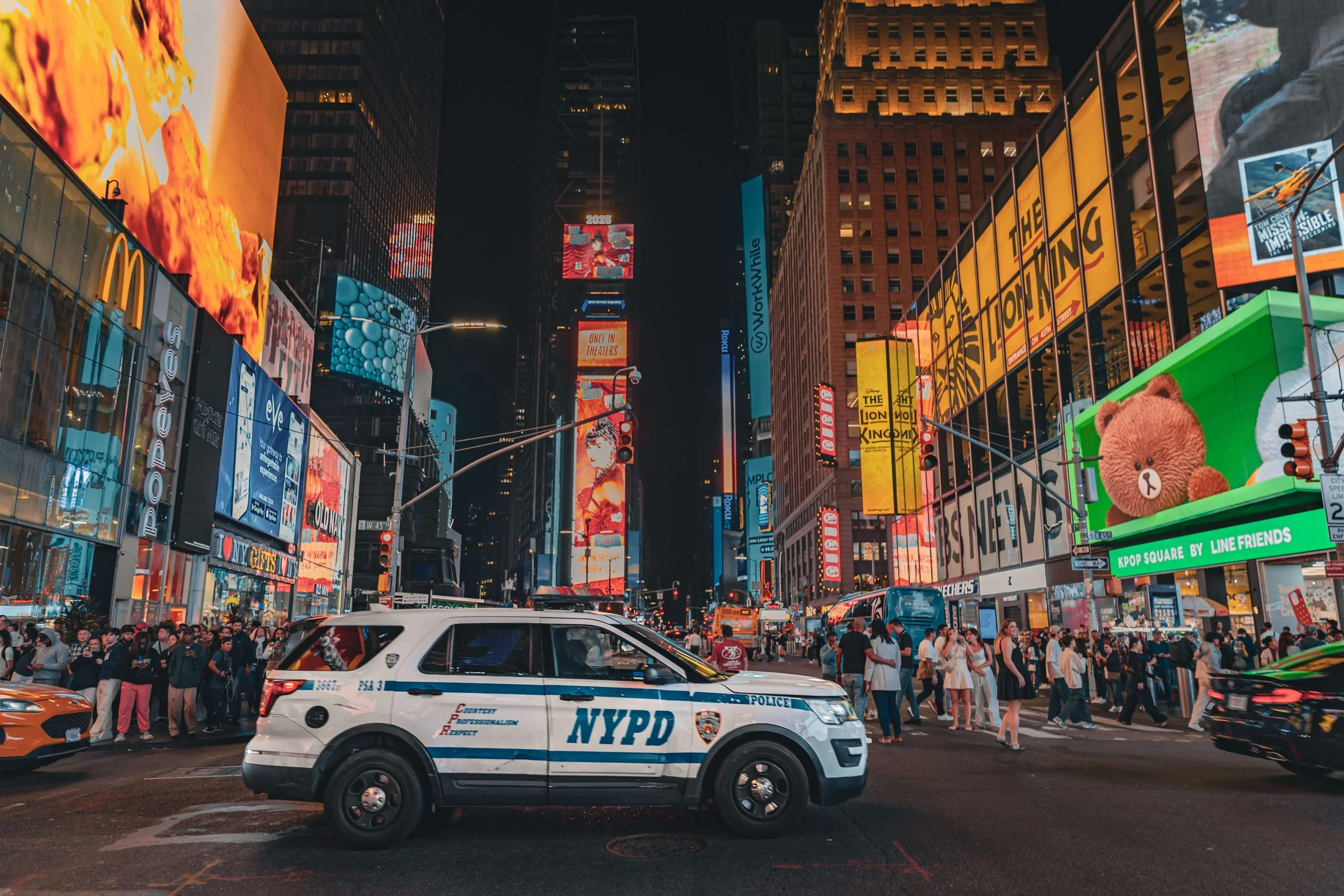 Nighttime scene in Times Square, New York City, with bright billboards and electronic advertisements, a crowd of pedestrians crossing the street, and an NYPD police vehicle in the foreground.