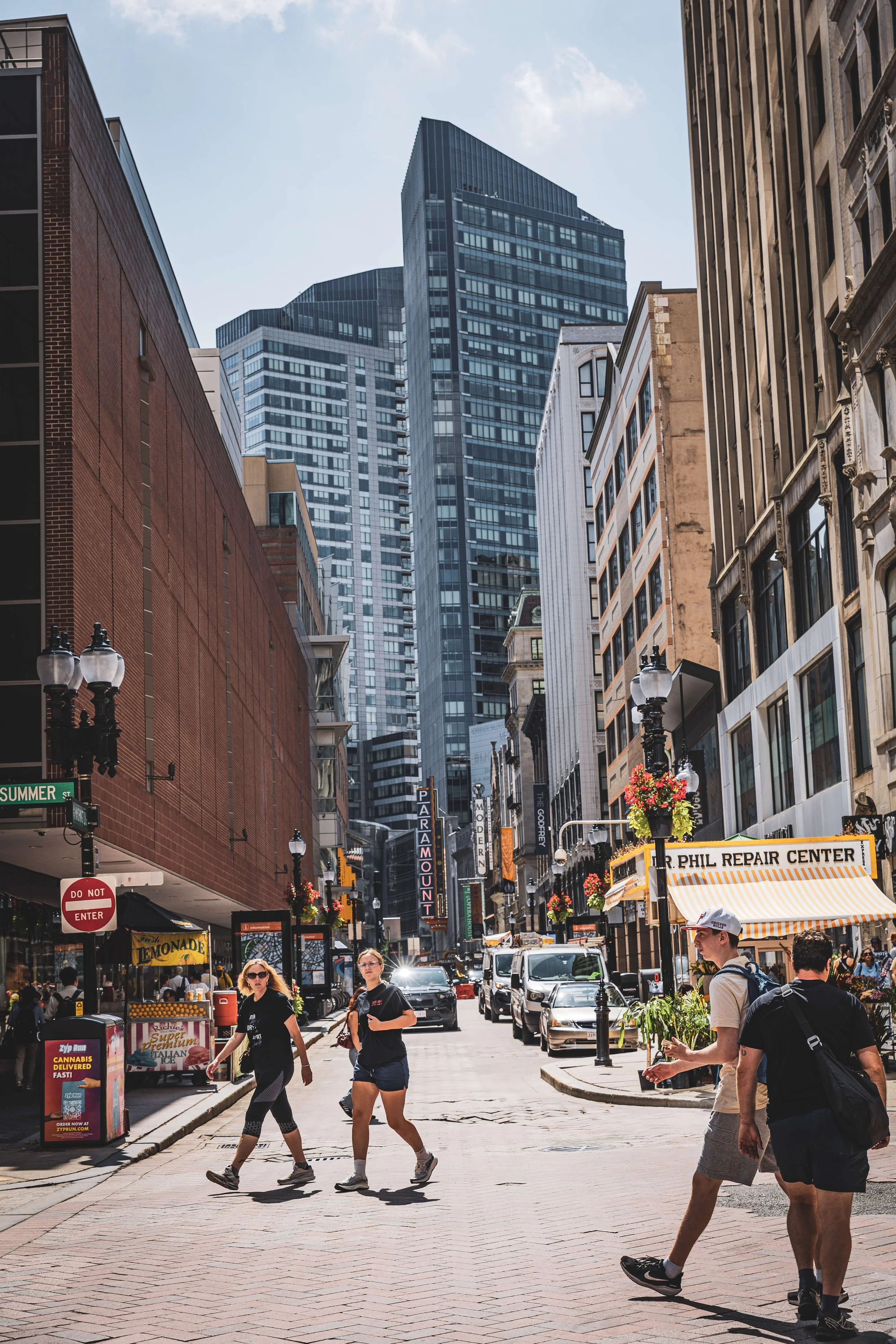 People walking on a busy city street with tall skyscrapers and shops, including a repair center and a market stall, under a cloudy sky.
