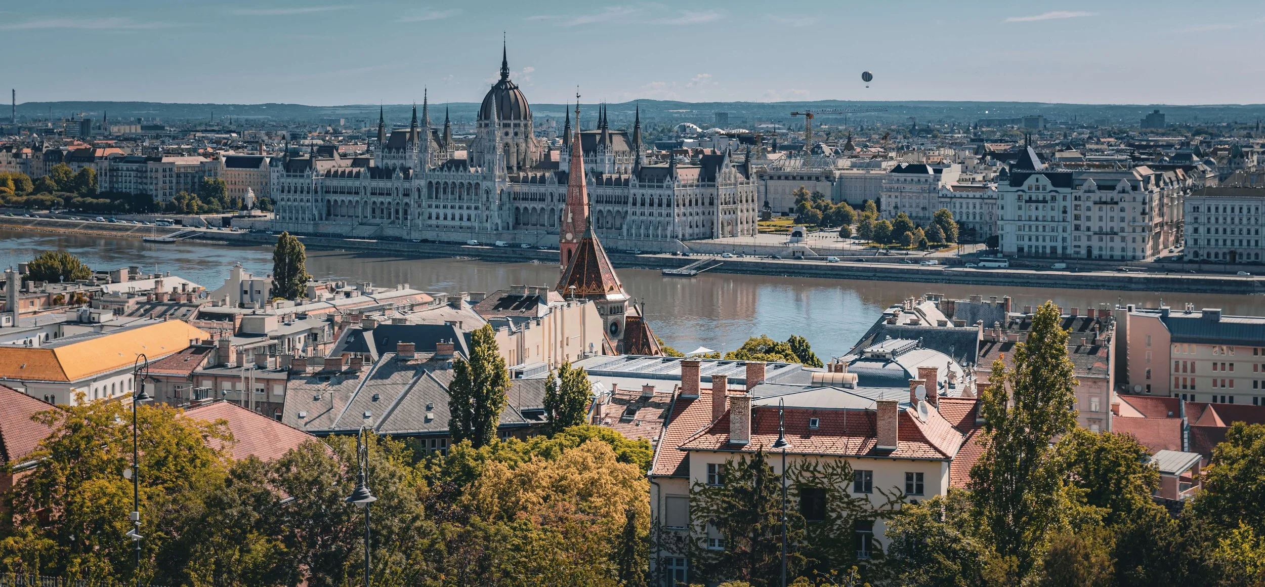 Panoramic view of Budapest, Hungary, featuring the Hungarian Parliament Building along the Danube River, with rooftops, trees, and a hot air balloon in the sky.
