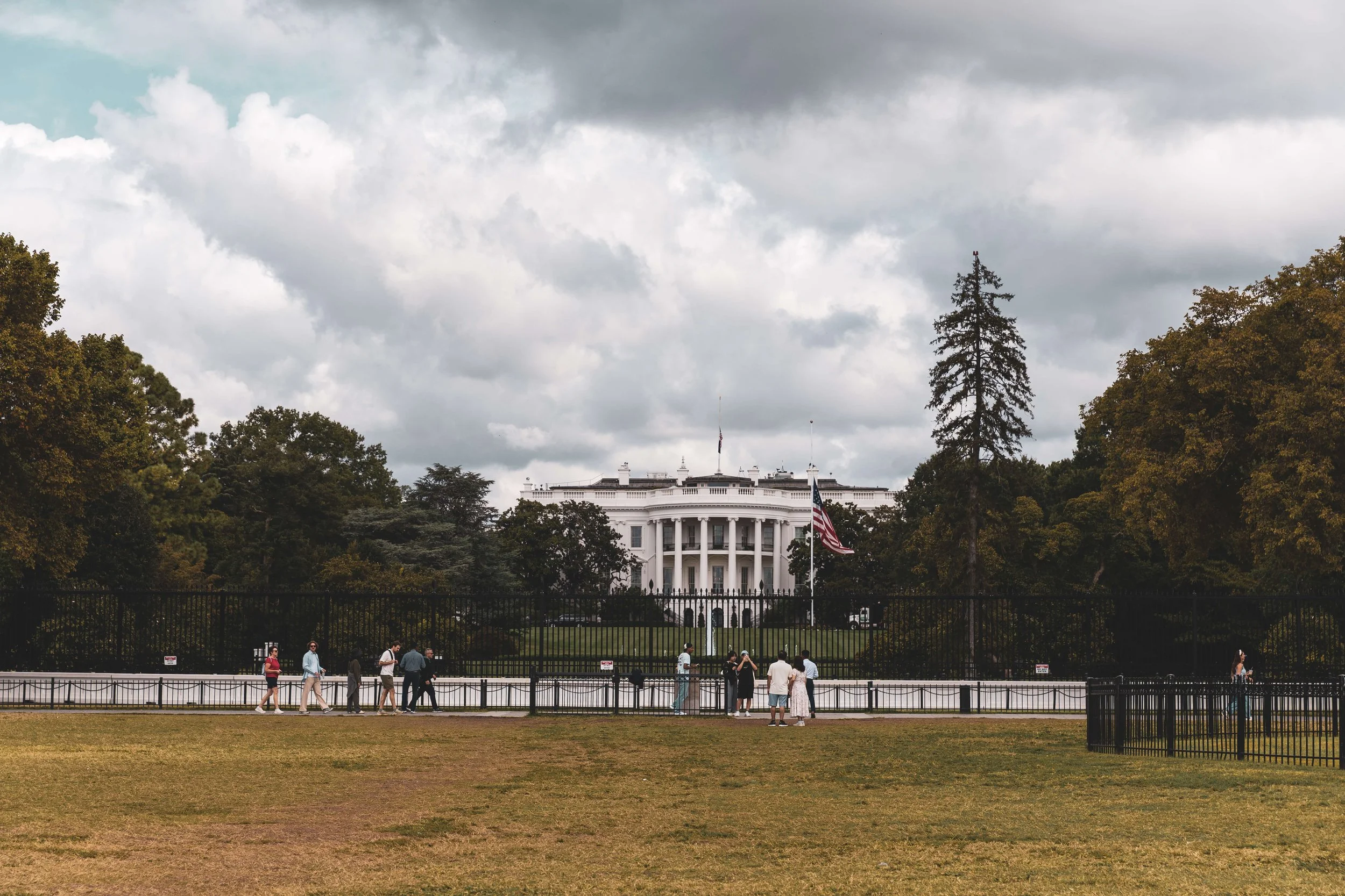 The White House in Washington D.C., seen through a fence with visitors walking in the foreground and cloudy skies overhead.