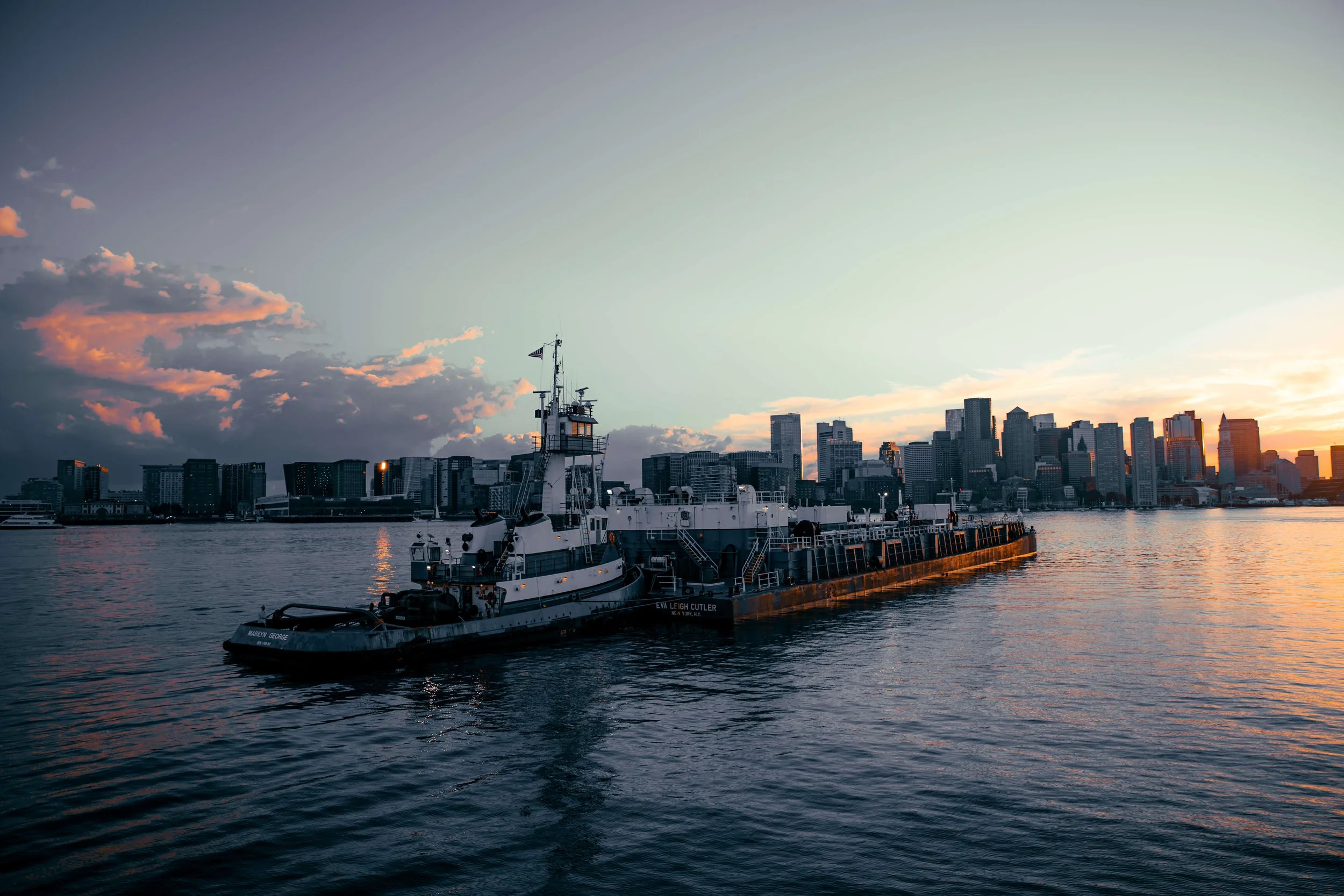 A tugboat named Eva Leigh Cutler on the water with a city skyline in the background during sunset.