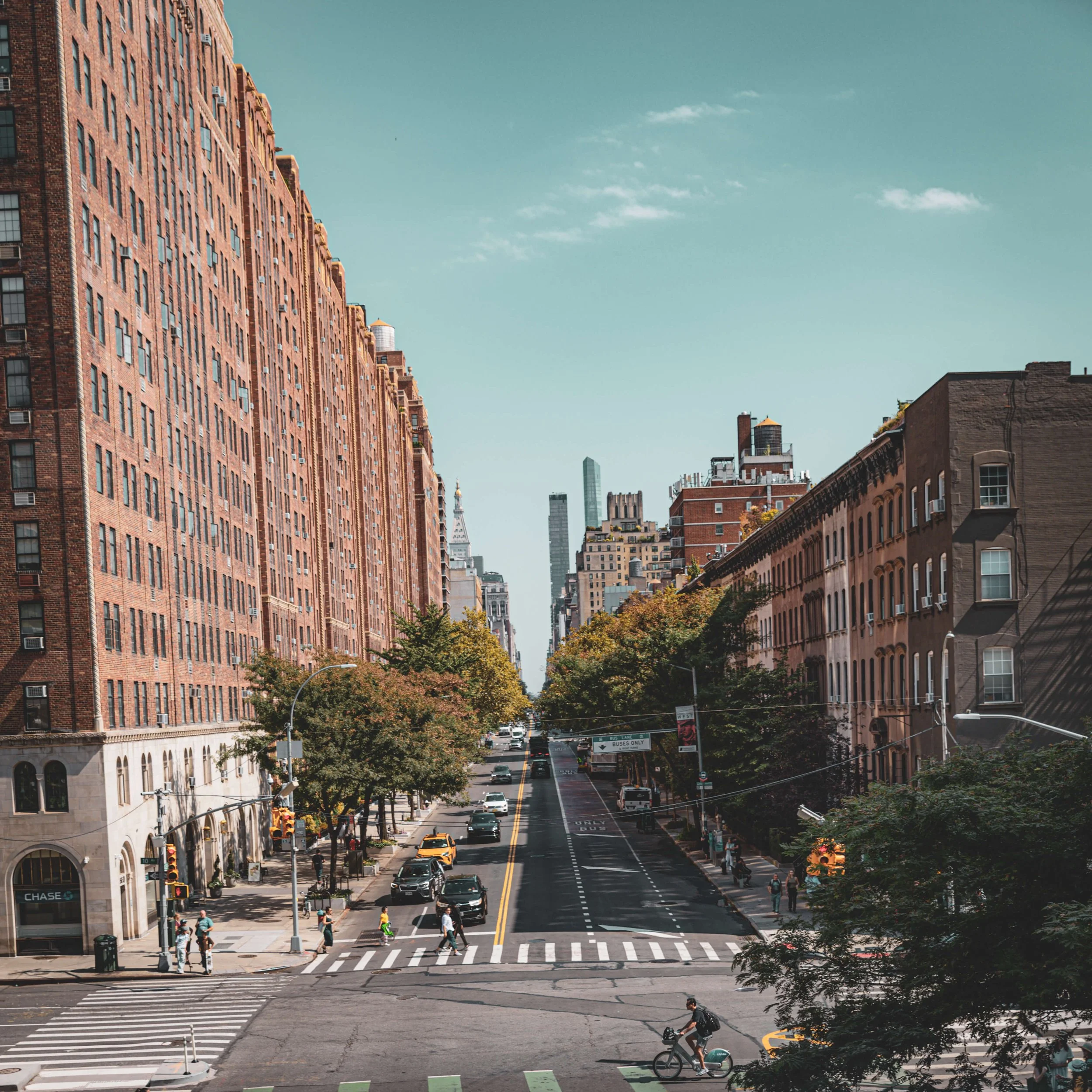 City street with tall brick buildings on both sides, trees lining the sidewalks, cars and pedestrians crossing the street on a sunny day.