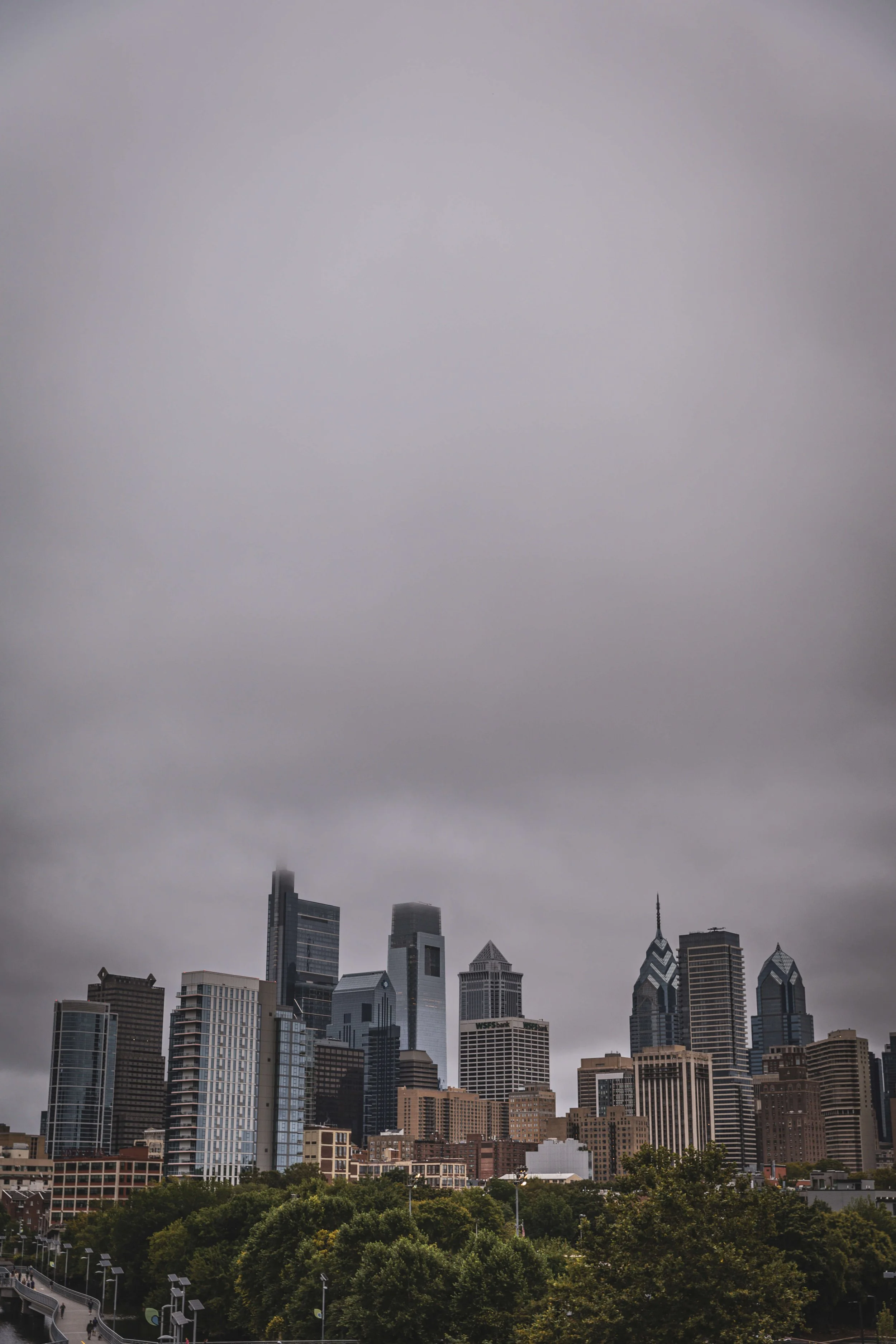 Skyline of a city with tall skyscrapers under a gray, overcast sky, and trees at the bottom.