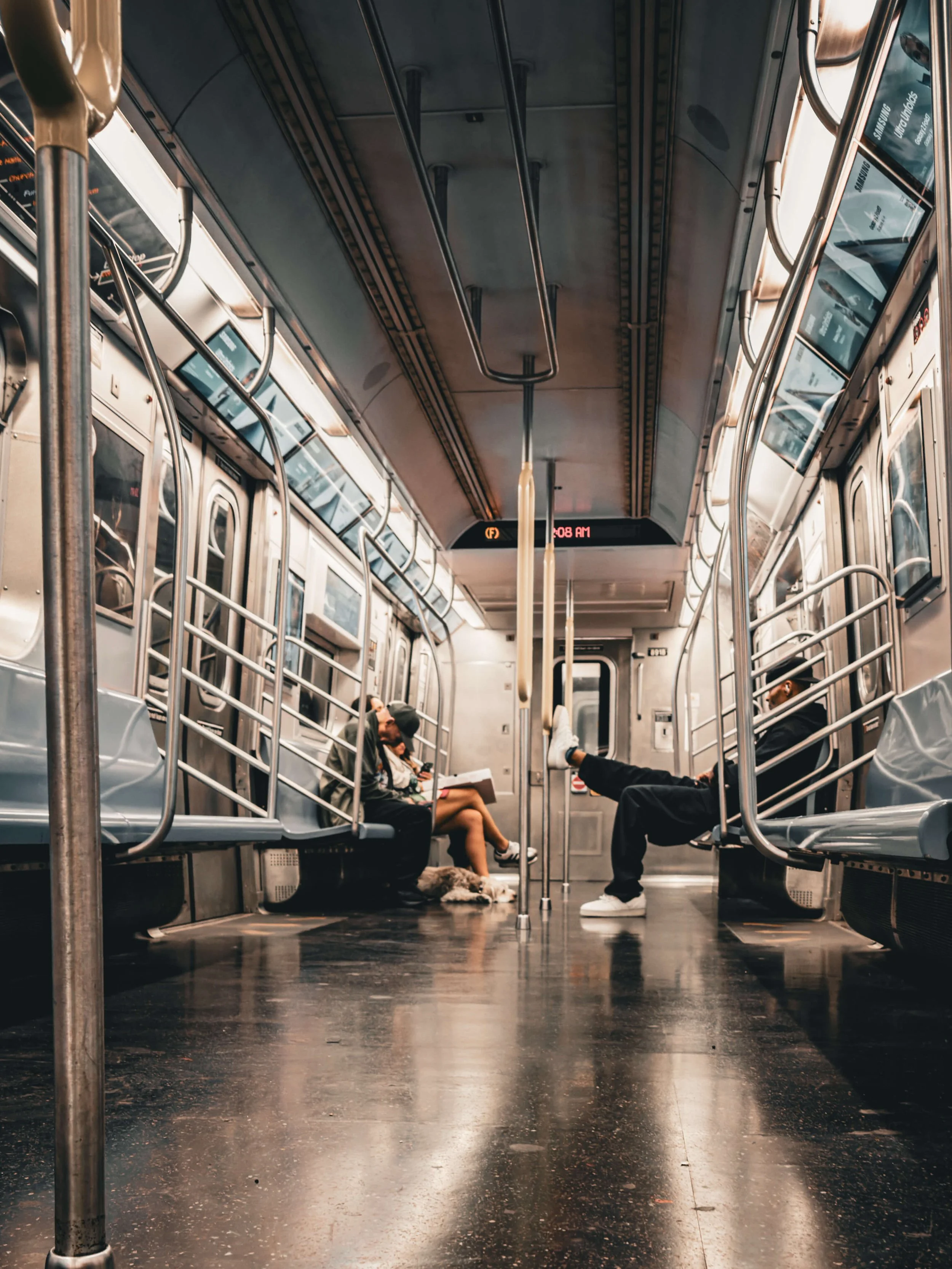 Inside a mostly empty subway car with two passengers seated, one man with his feet up and another person reading, and a dog lying on the floor.