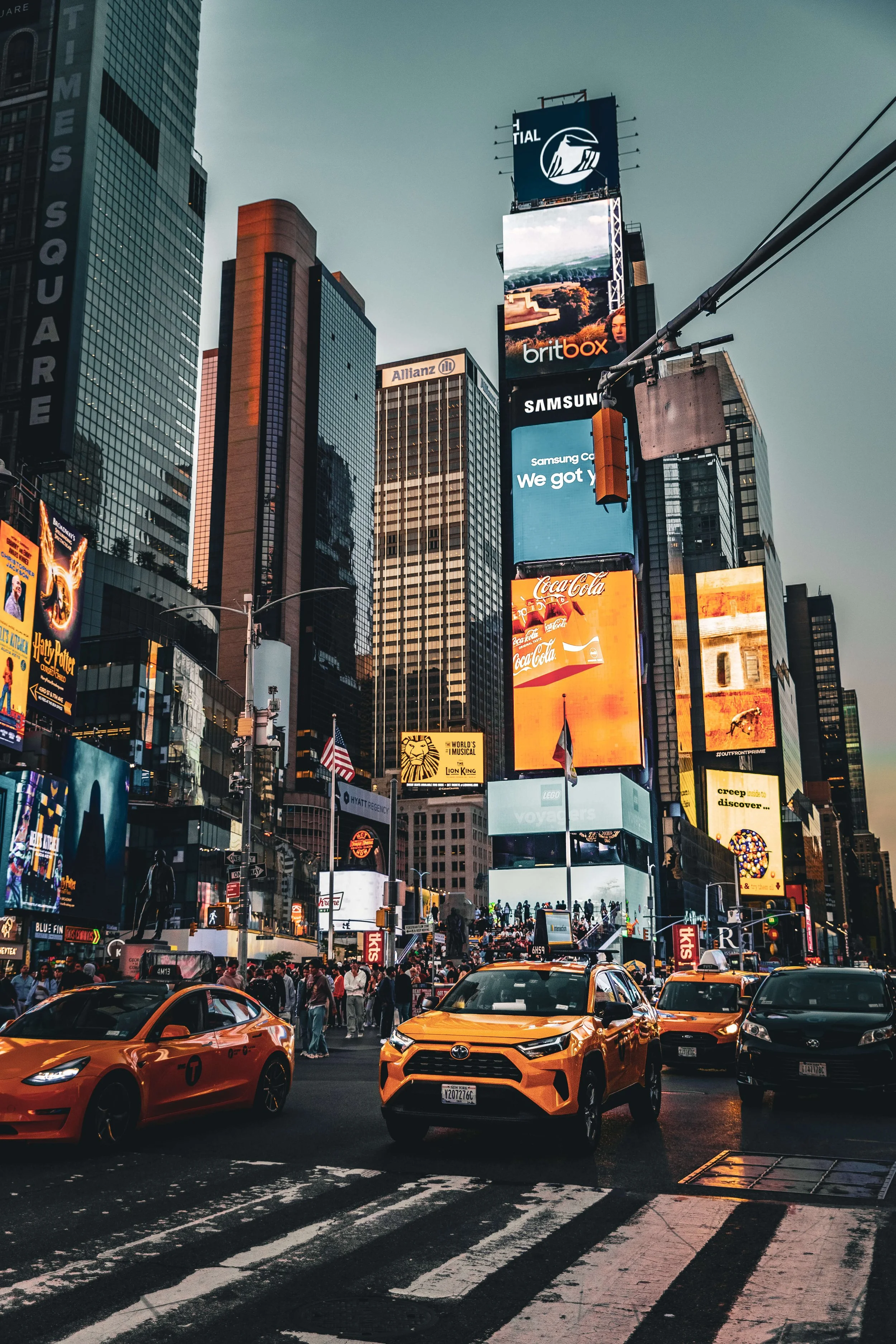 Times Square in New York City showing tall skyscrapers, large digital billboards, yellow taxis, and a busy crowd crossing the street.
