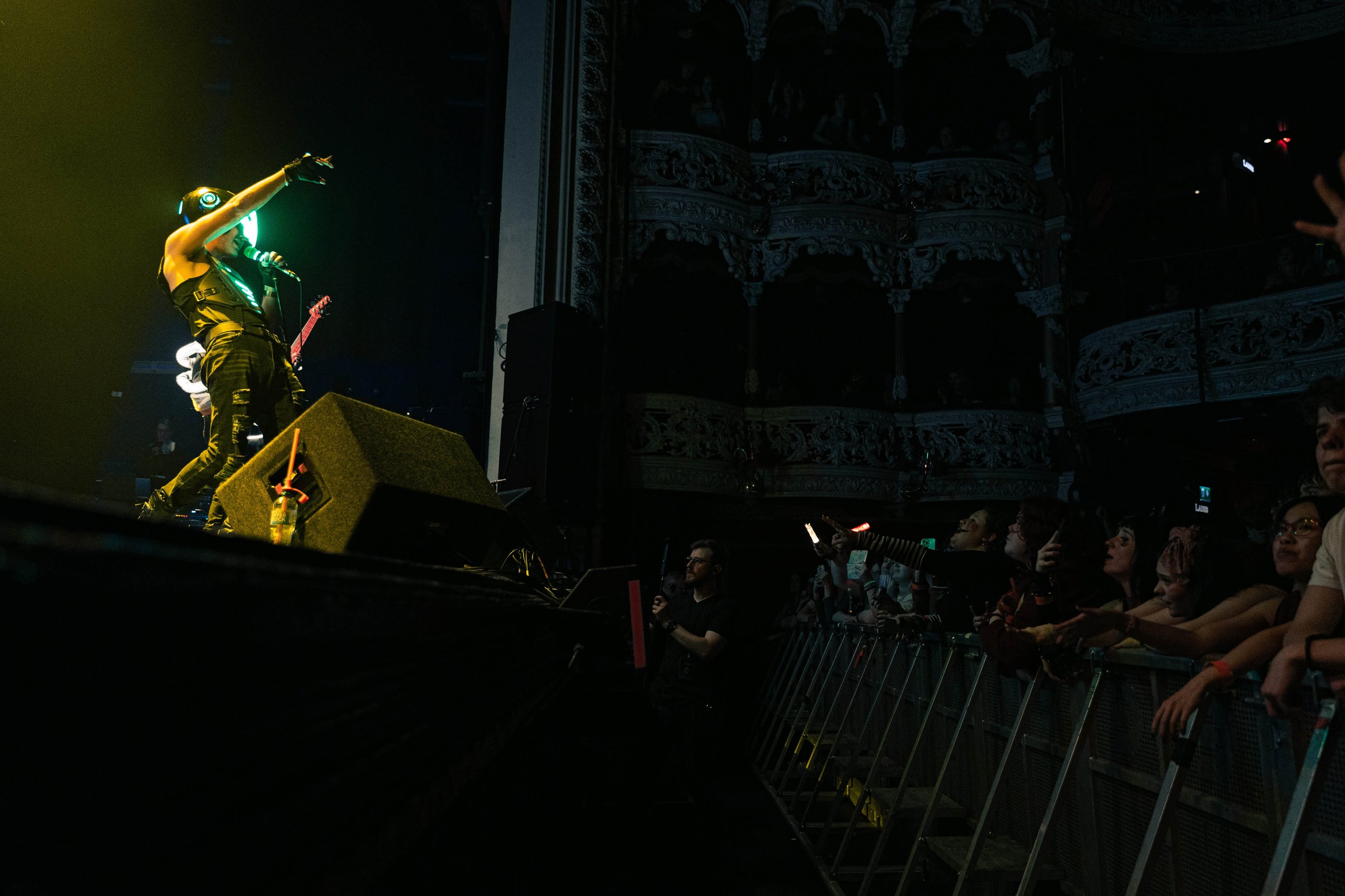 Performer on stage wearing a virtual reality headset, holding a microphone, with an audience watching in a dimly lit theater.