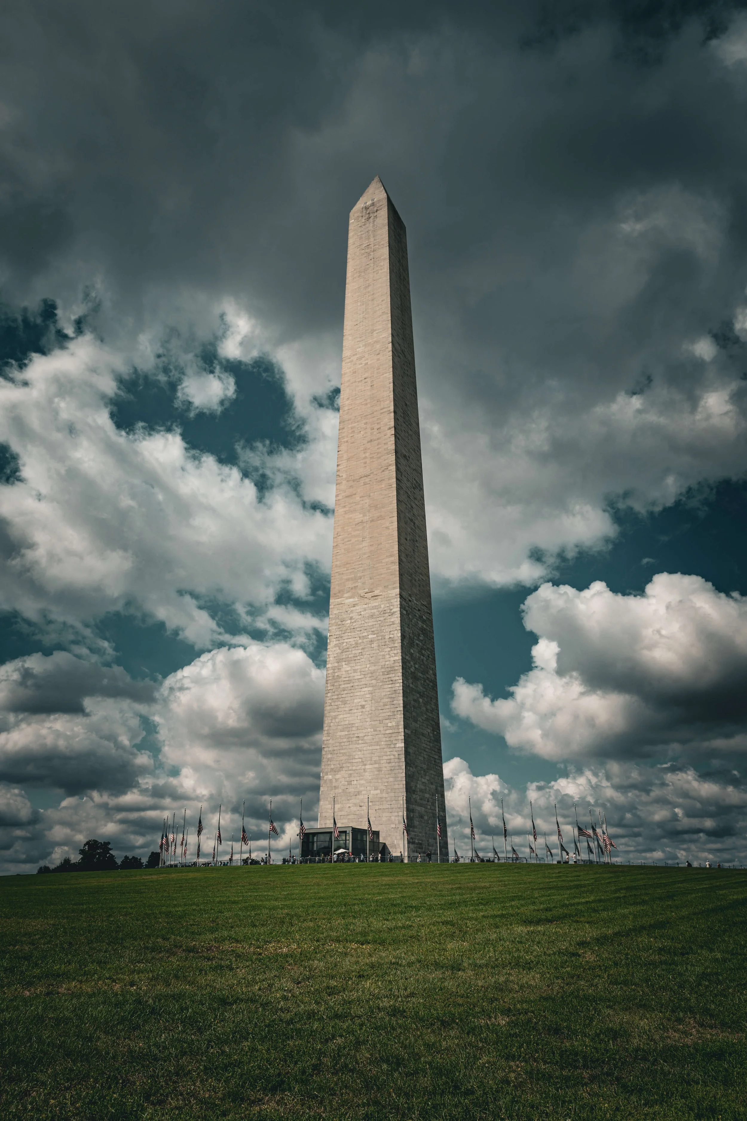 The Washington Monument, an obelisk in Washington D.C., surrounded by American flags, with a cloudy sky overhead and a grassy area in the foreground.