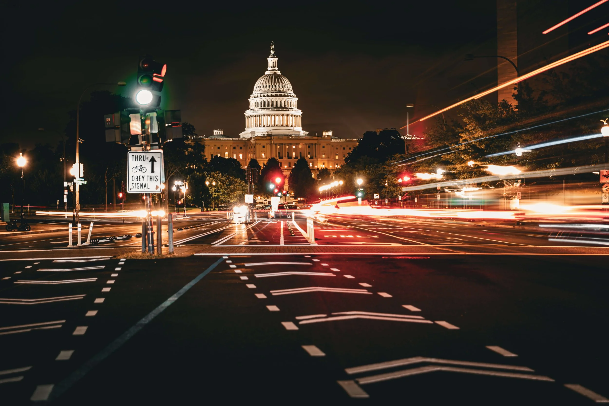 Nighttime view of the U.S. Capitol building in Washington, D.C., with light trails from moving vehicles, traffic lights, and a sign instructing to obey signals for bicycles.