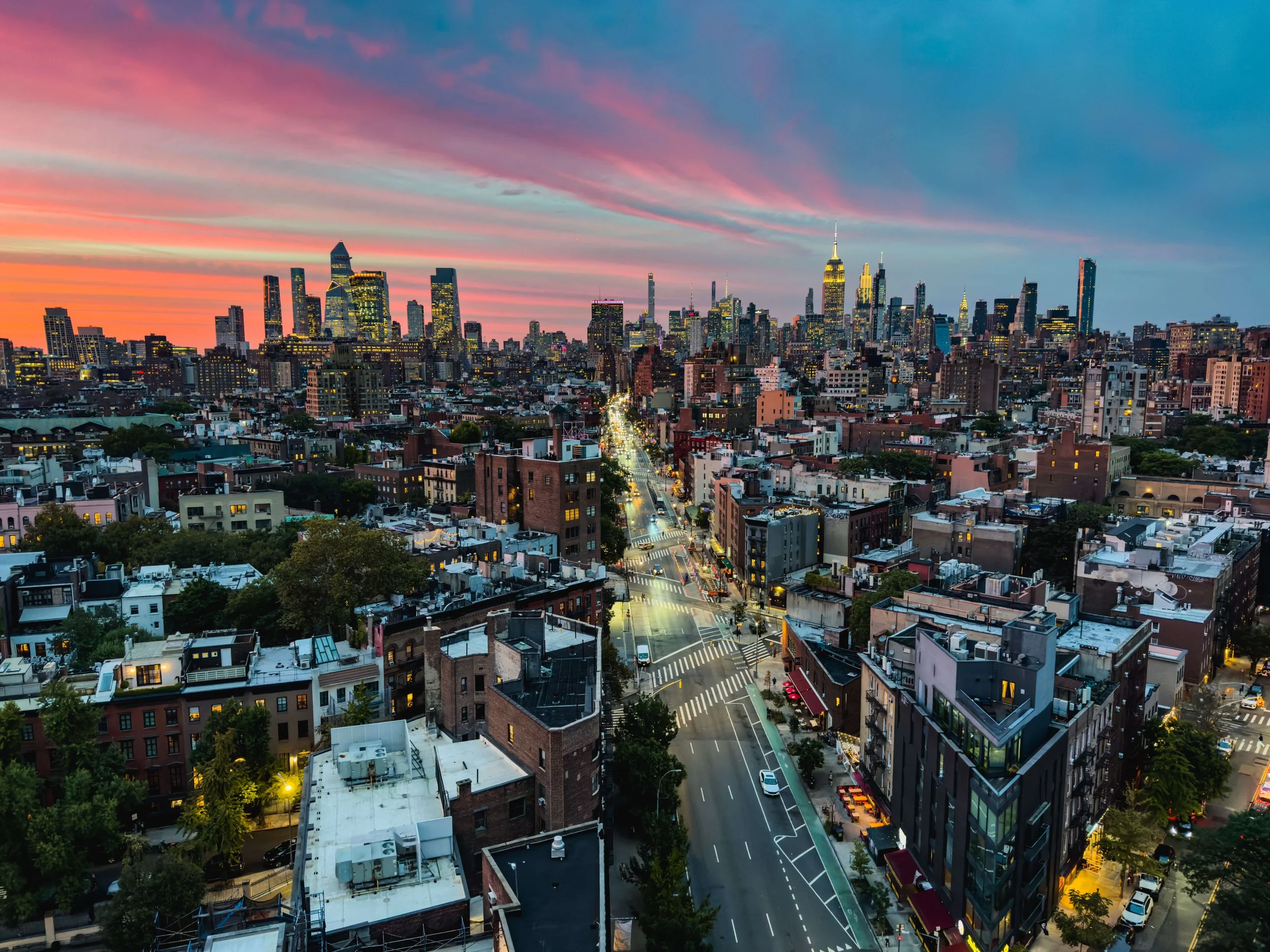 Sunset over New York City skyline with colorful sky, tall buildings, and a busy street in the foreground.