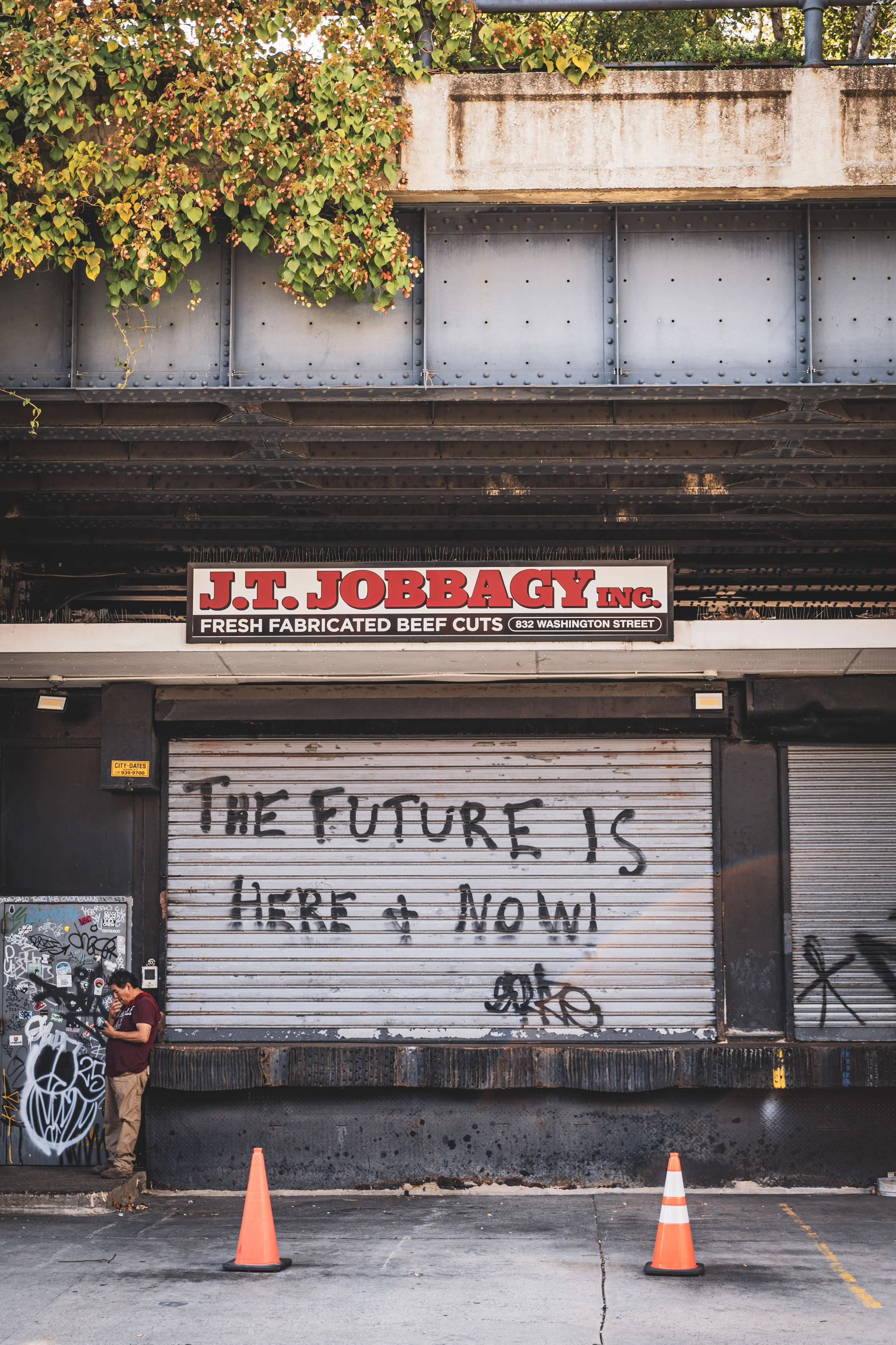 Urban storefront with graffiti and a sign for J.T. Jobbagy Inc. fresh fabricated beef cuts, and a large spray-painted message on the shutter that reads 'The future is here & now.'