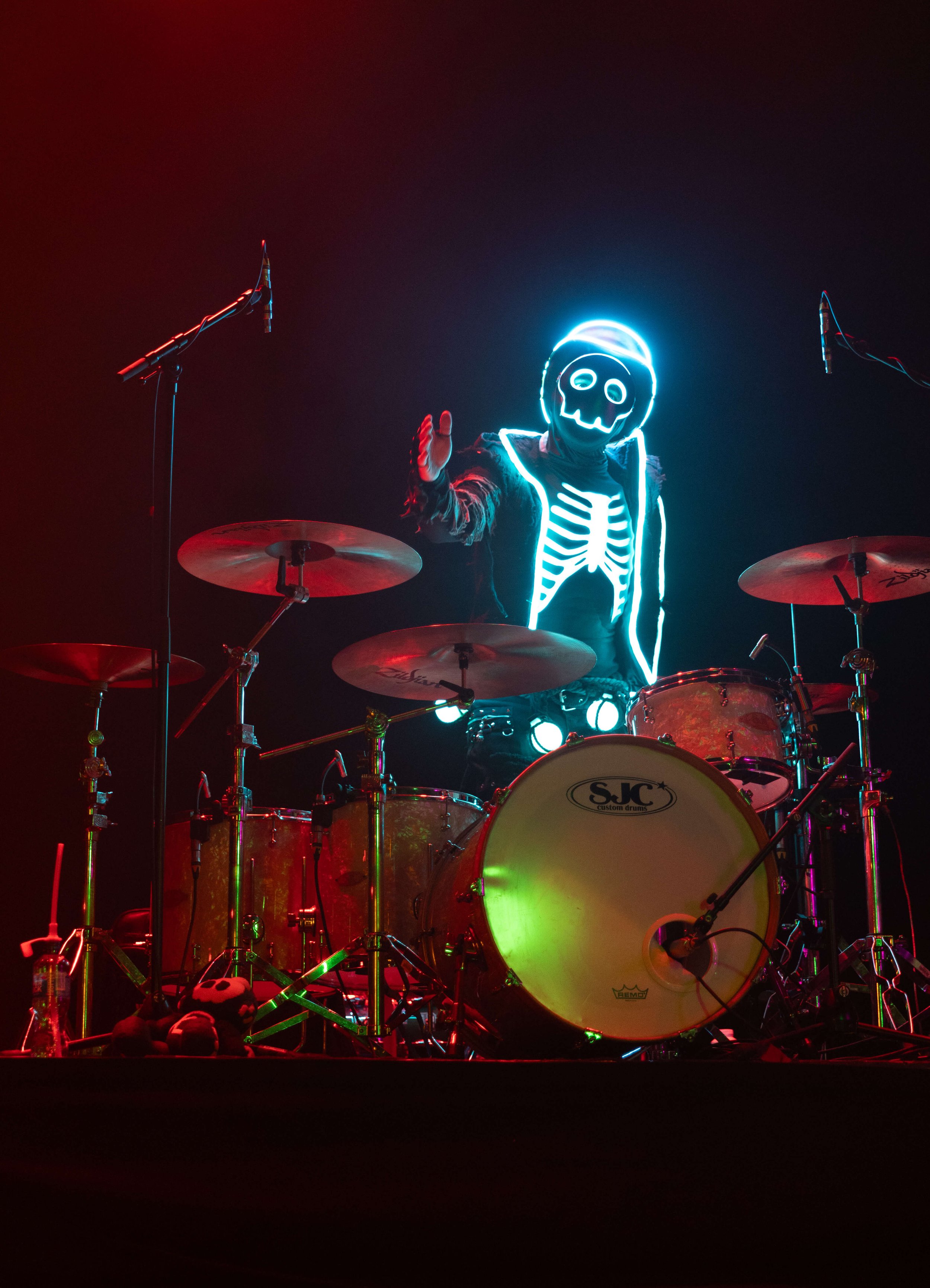 Person wearing a glowing skeleton costume with a skull mask, playing a drum set on stage at night with microphones overhead.