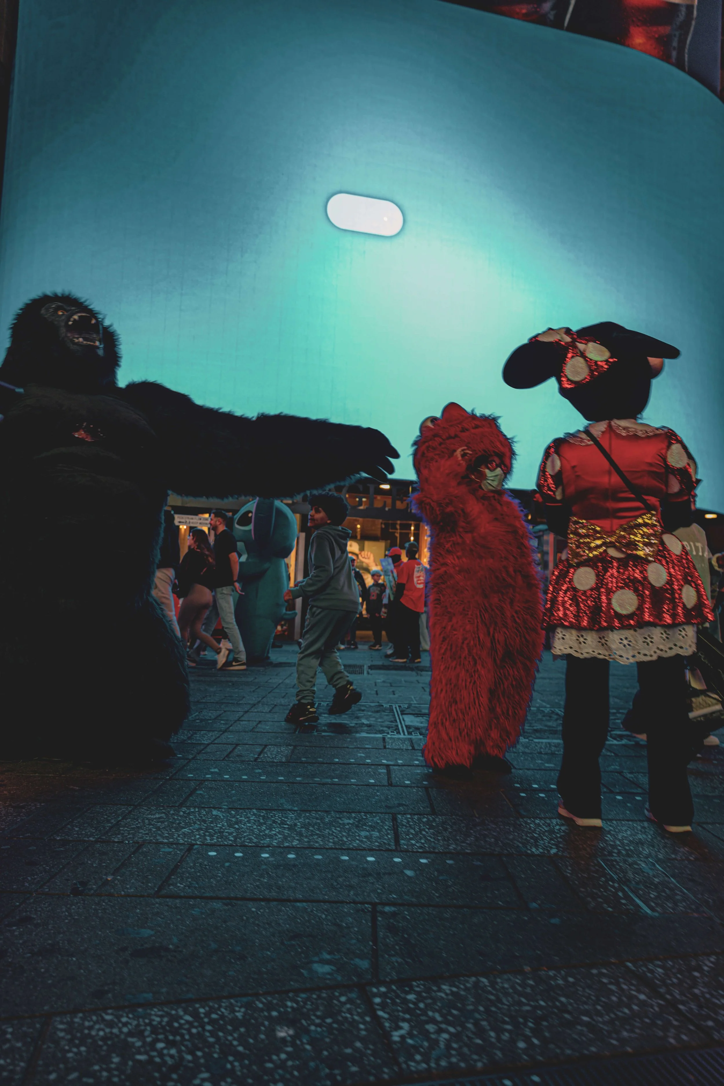 People at a Halloween event are dressed in costumes resembling a gorilla, a red furry monster, and a ladybug dress, with a large LED screen in the background.