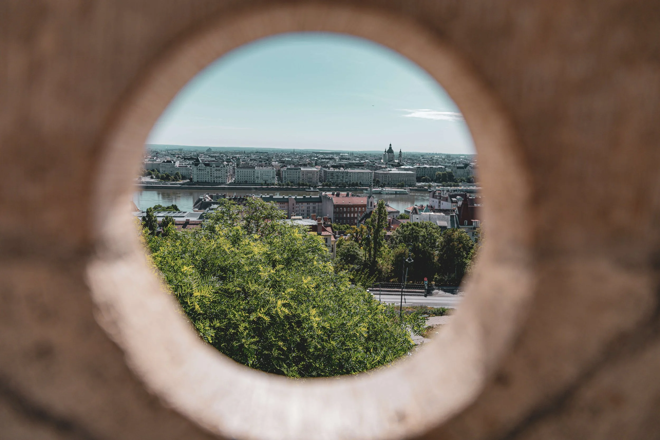 Cityscape view of a river with buildings and a church steeple, seen through a circular opening in a brick or stone structure.