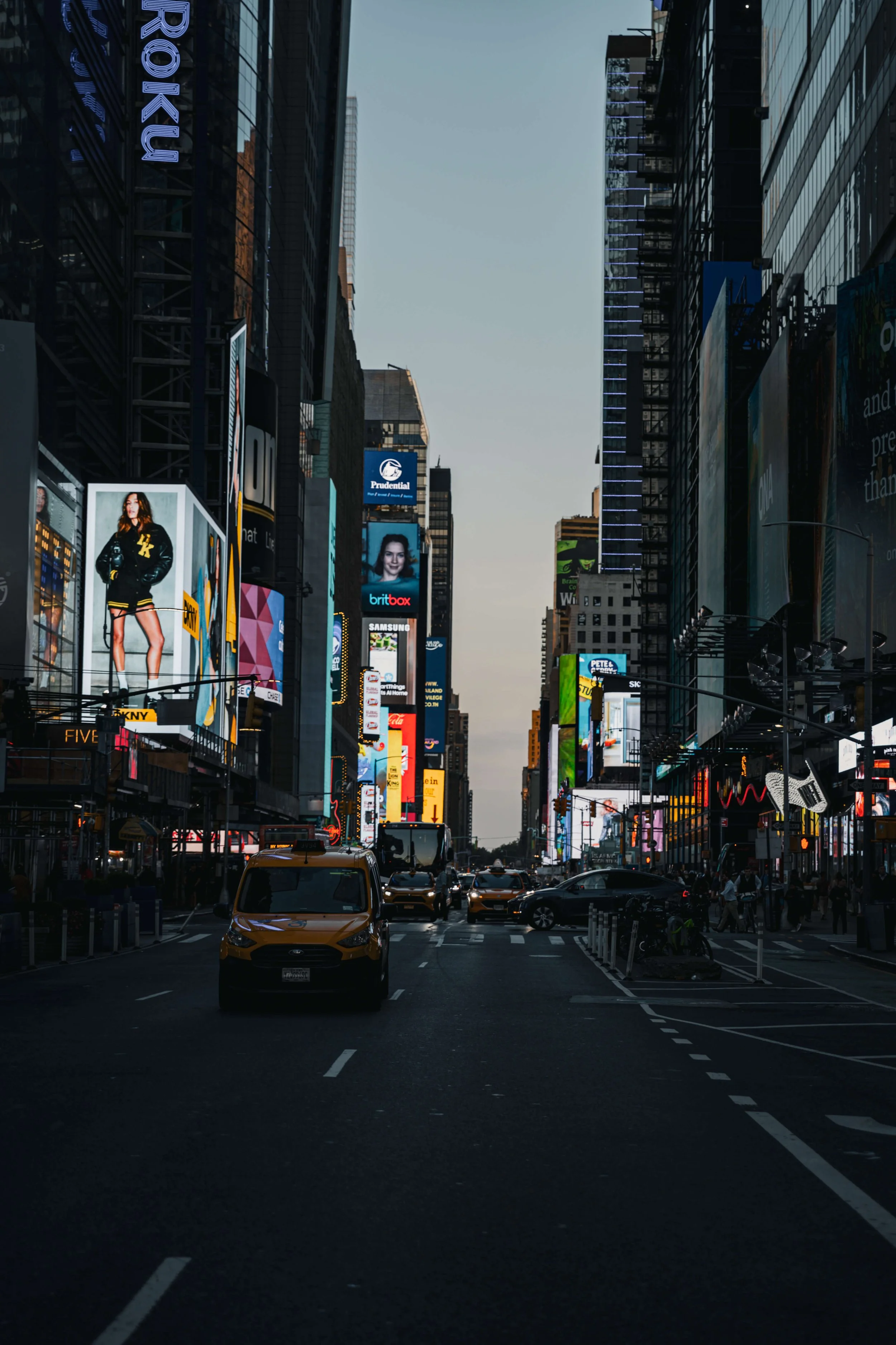 View of Times Square in New York City at dusk, featuring illuminated billboards and yellow taxis on the street.
