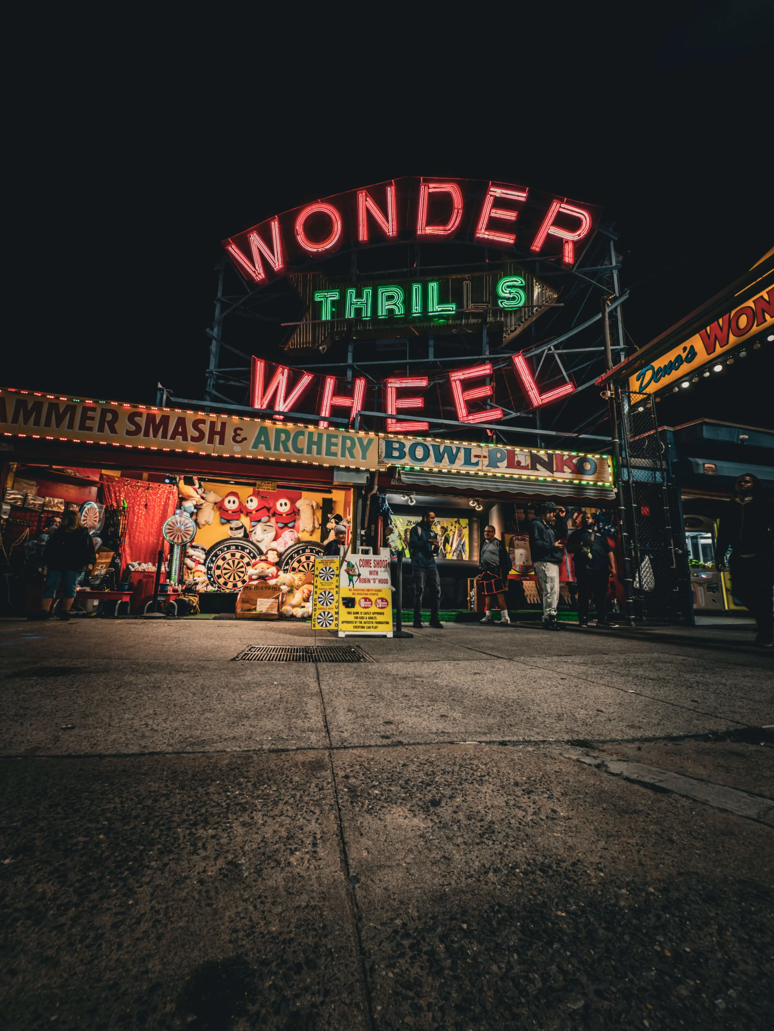 Nighttime scene of a fair with illuminated neon signs reading 'Wonder Wheel' and 'Thrill's.' There are booths and people walking around, with games and prizes visible on the left side.