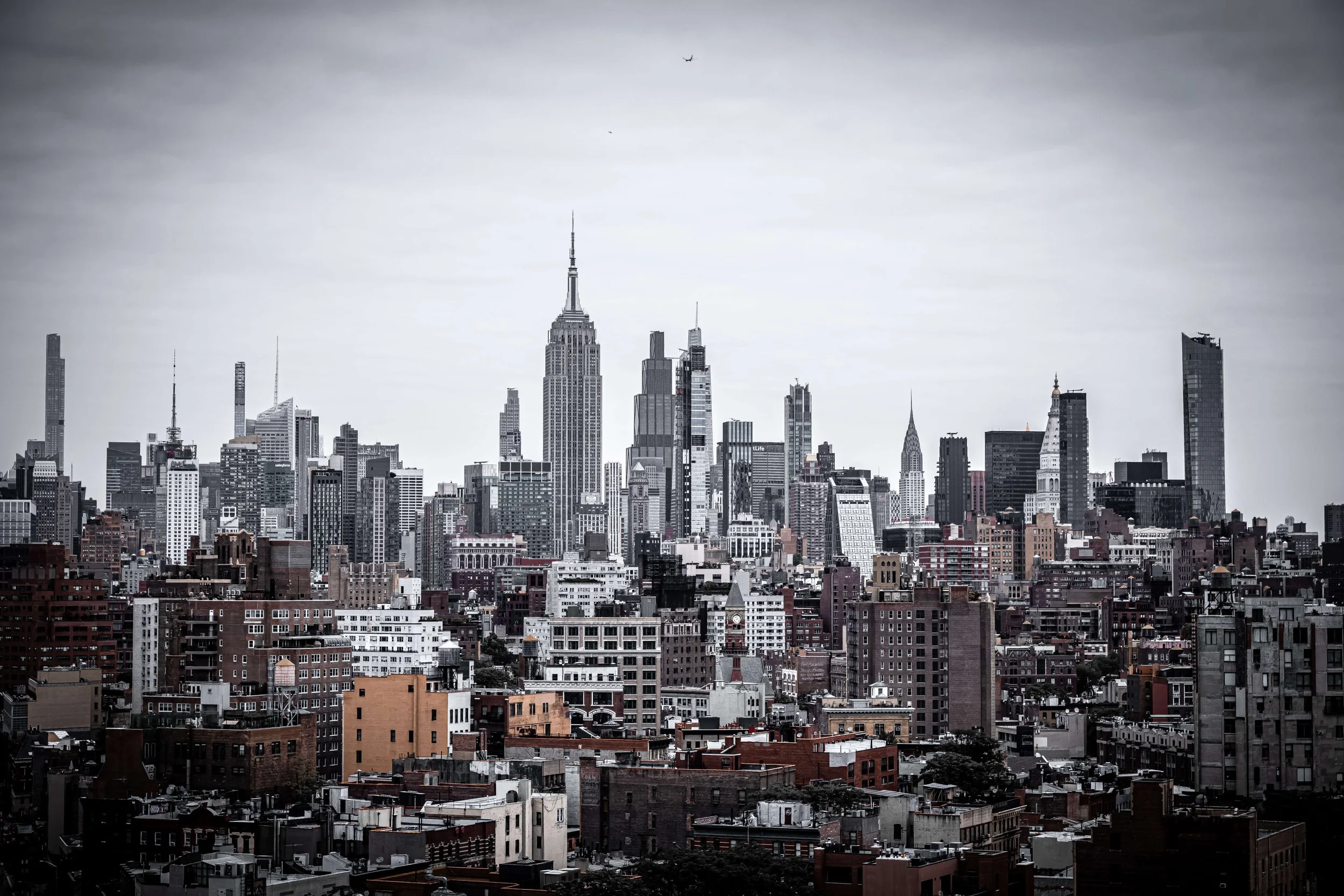 Black and white cityscape of Manhattan with tall skyscrapers, including the Empire State Building, under a cloudy sky.