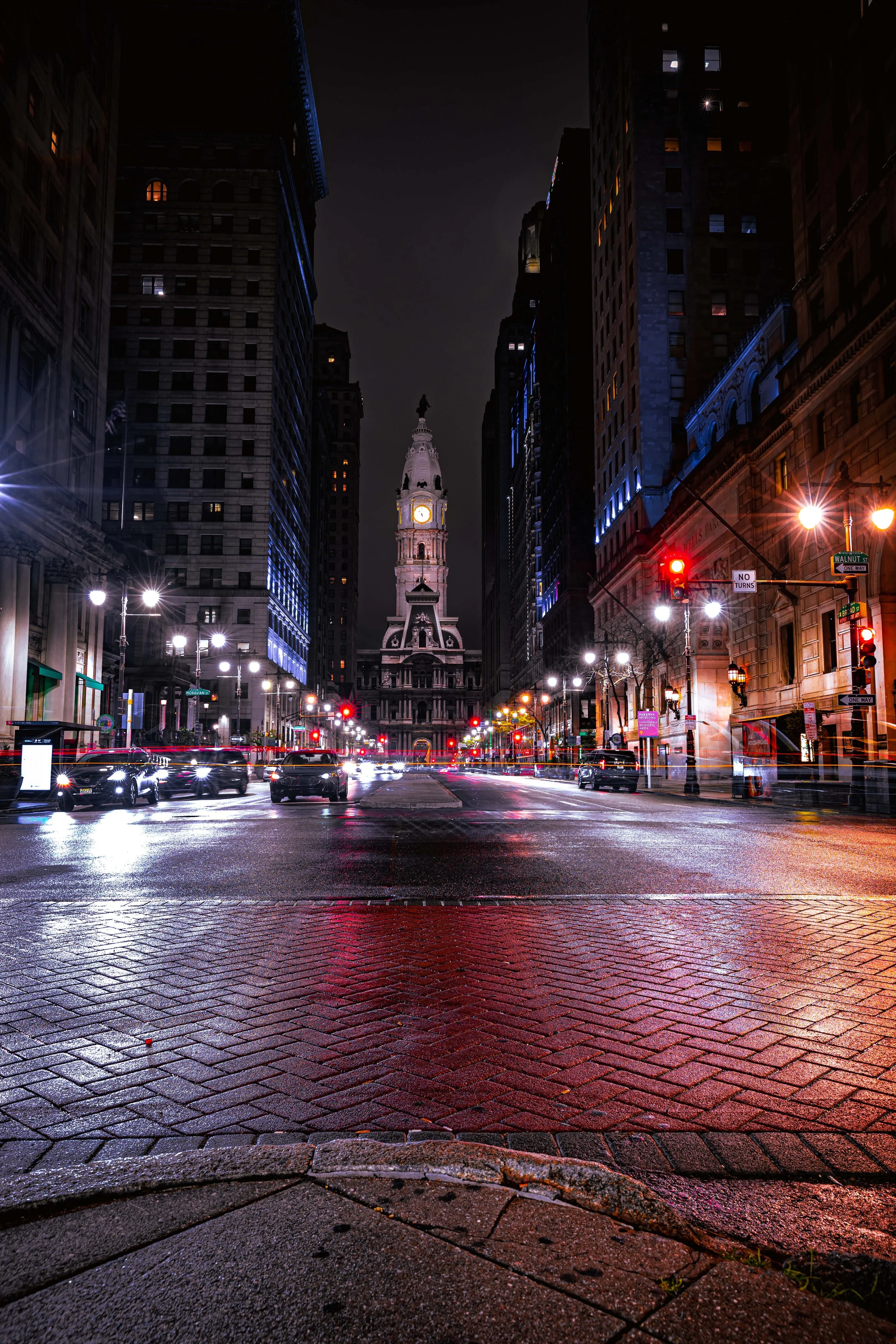 Nighttime view of a city street with a historic clock tower in the distance, bright streetlights, blurred car lights, and tall buildings on both sides.