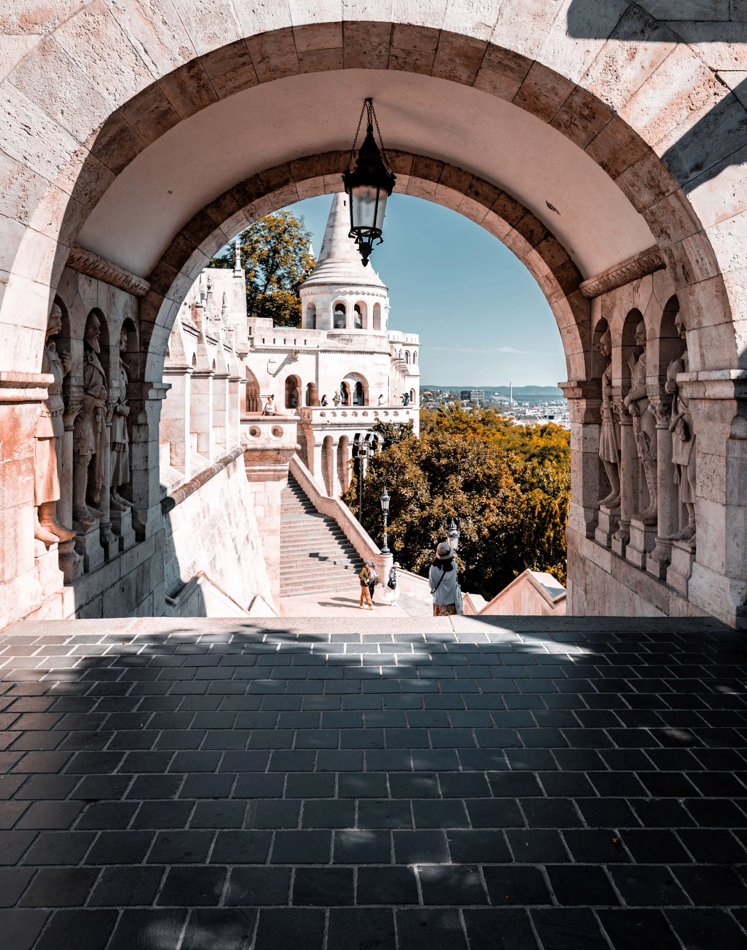 View of Fisherman's Bastion in Budapest, Hungary through a stone archway, with a medieval-style tower and decorative statues visible in the background. The scene includes a pathway with people and trees with autumn foliage.