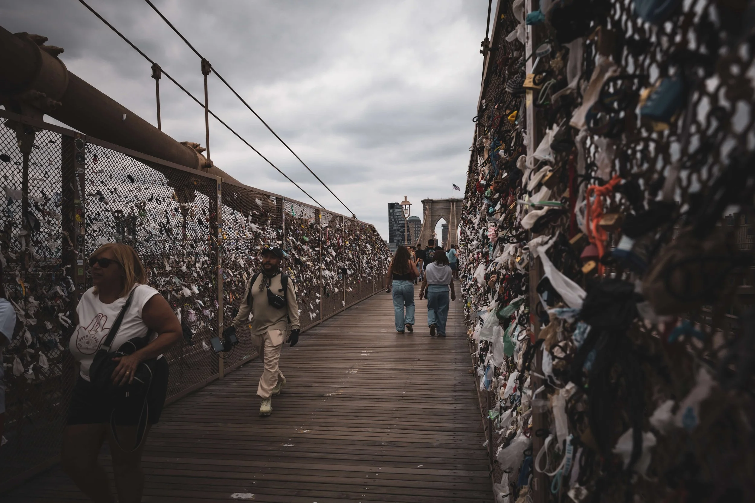 People walking on the Brooklyn Bridge with numerous padlocks attached to the fence on both sides.