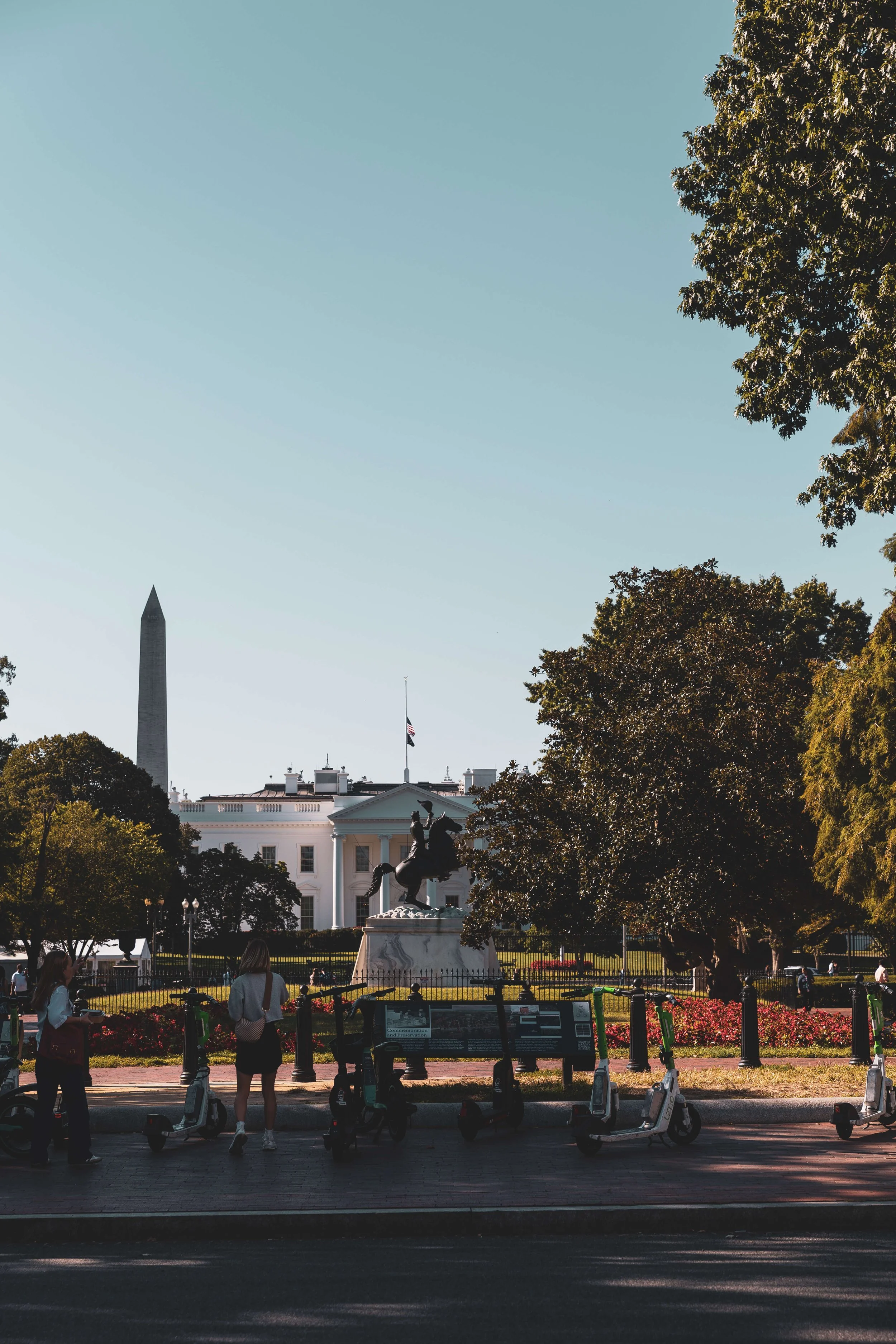 View of the White House with a statue in front, surrounded by trees and flowers, and the Washington Monument in the background.