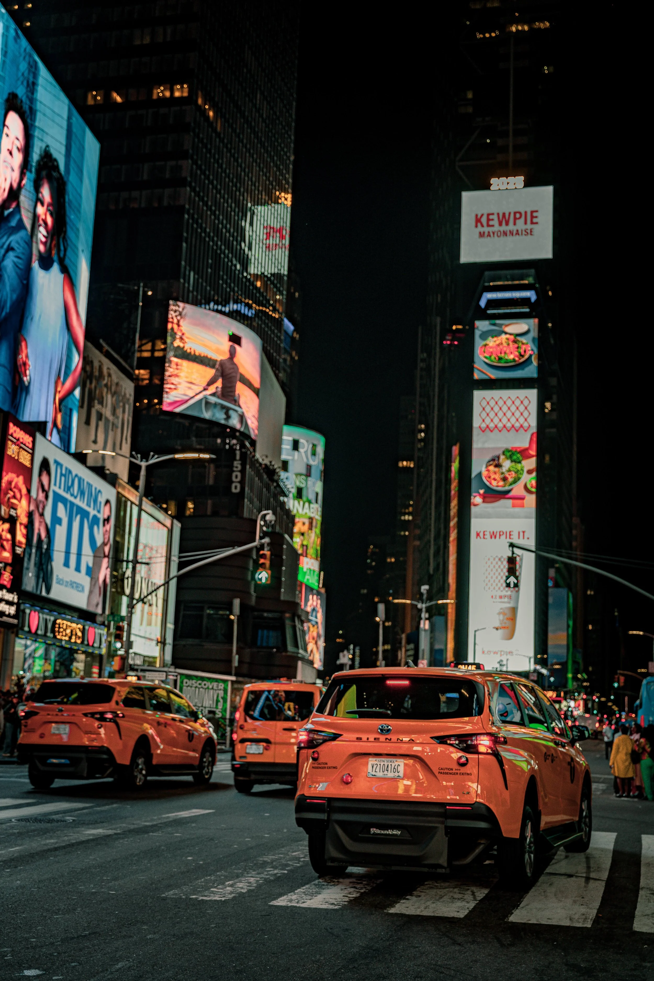 Nighttime scene of Times Square in New York City with bright digital billboards, advertisements, and taxis parked at a crosswalk.