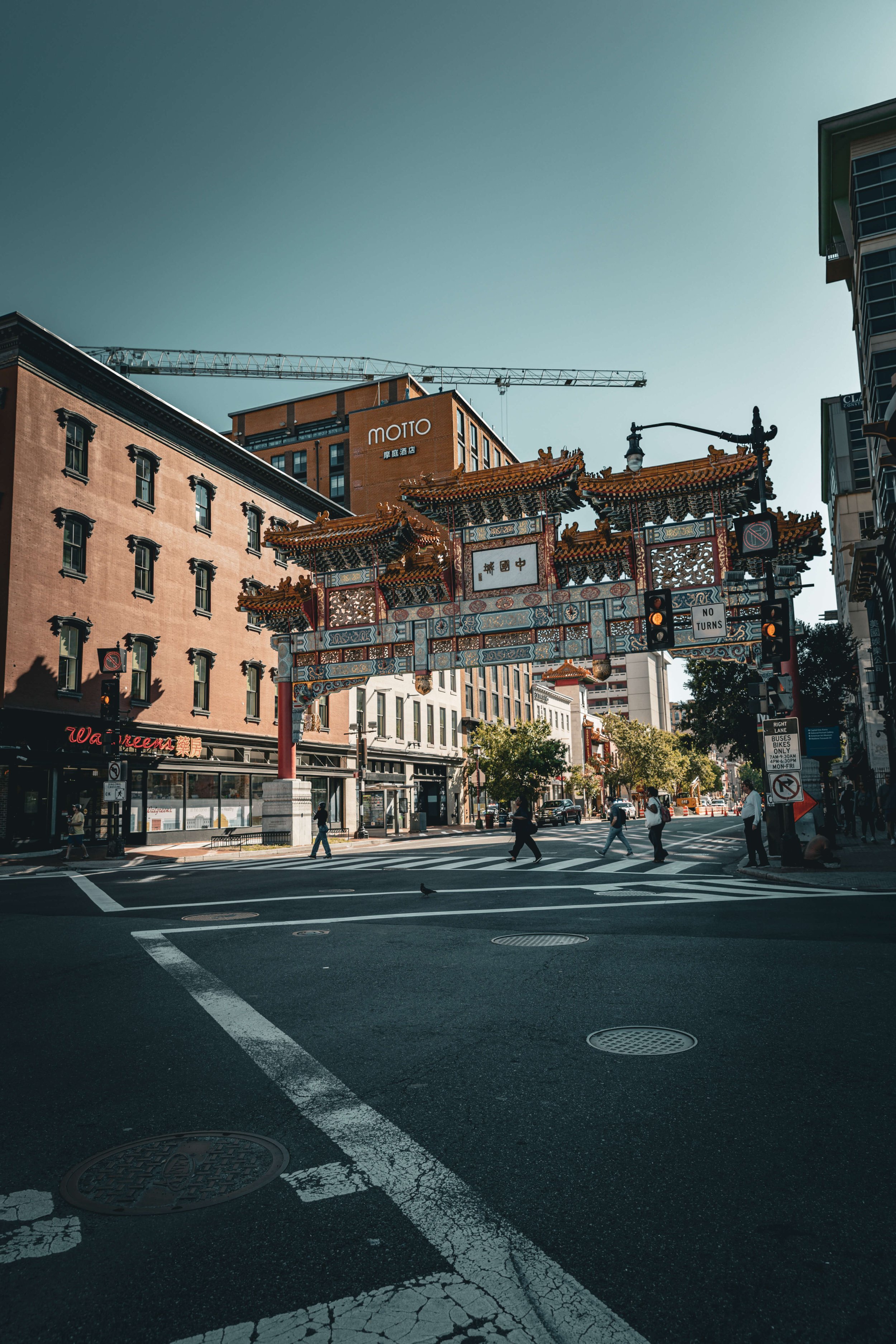 Urban street scene in Chinatown with a traditional Chinese archway over crosswalk, pedestrians crossing, surrounding modern buildings, and clear sky.