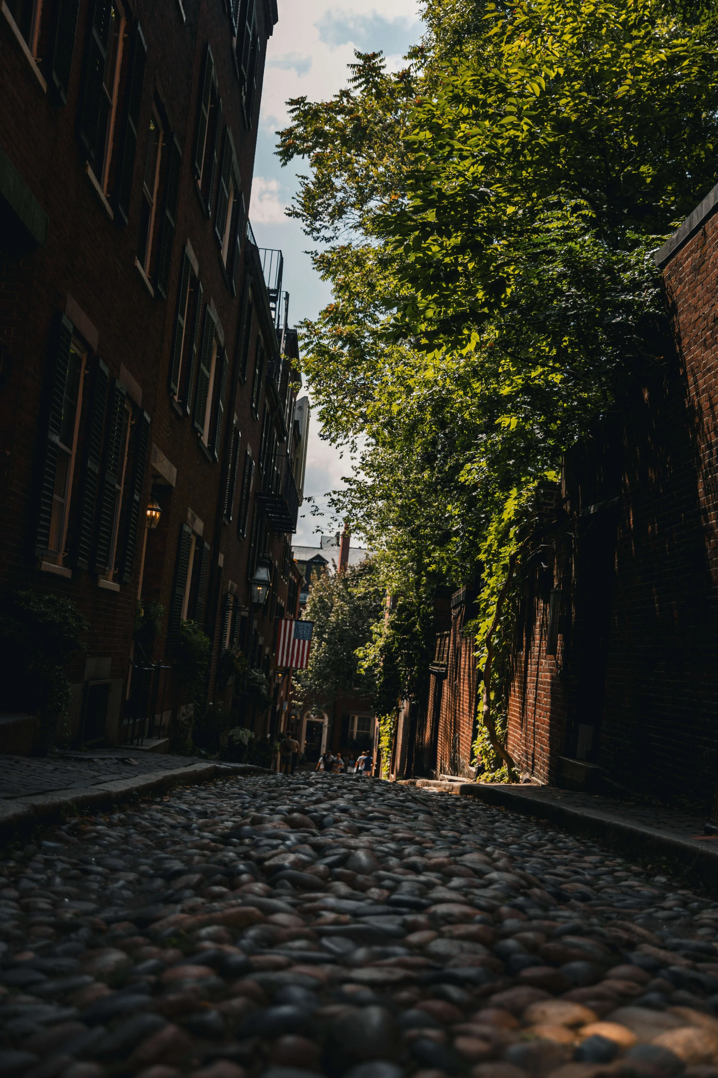 A cobblestone street in a historic city with brick buildings and green trees, with a flag hanging from one of the buildings and a small group of people in the distance.