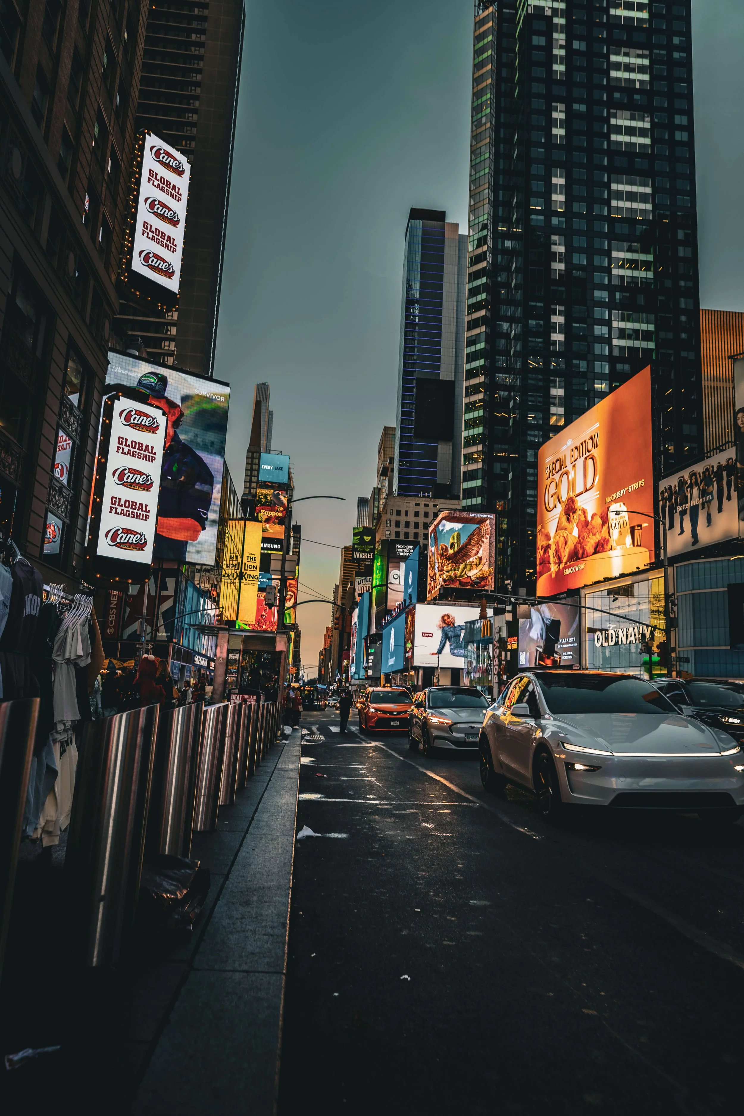 City street scene at dusk with tall buildings, digital billboards, and cars parked along the curb.