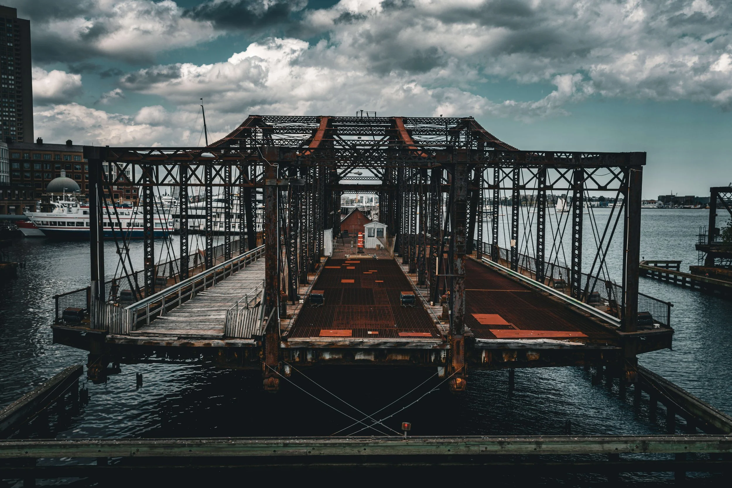Old rusty dock or structure extending over water with boats docked on the side, cloudy sky above.
