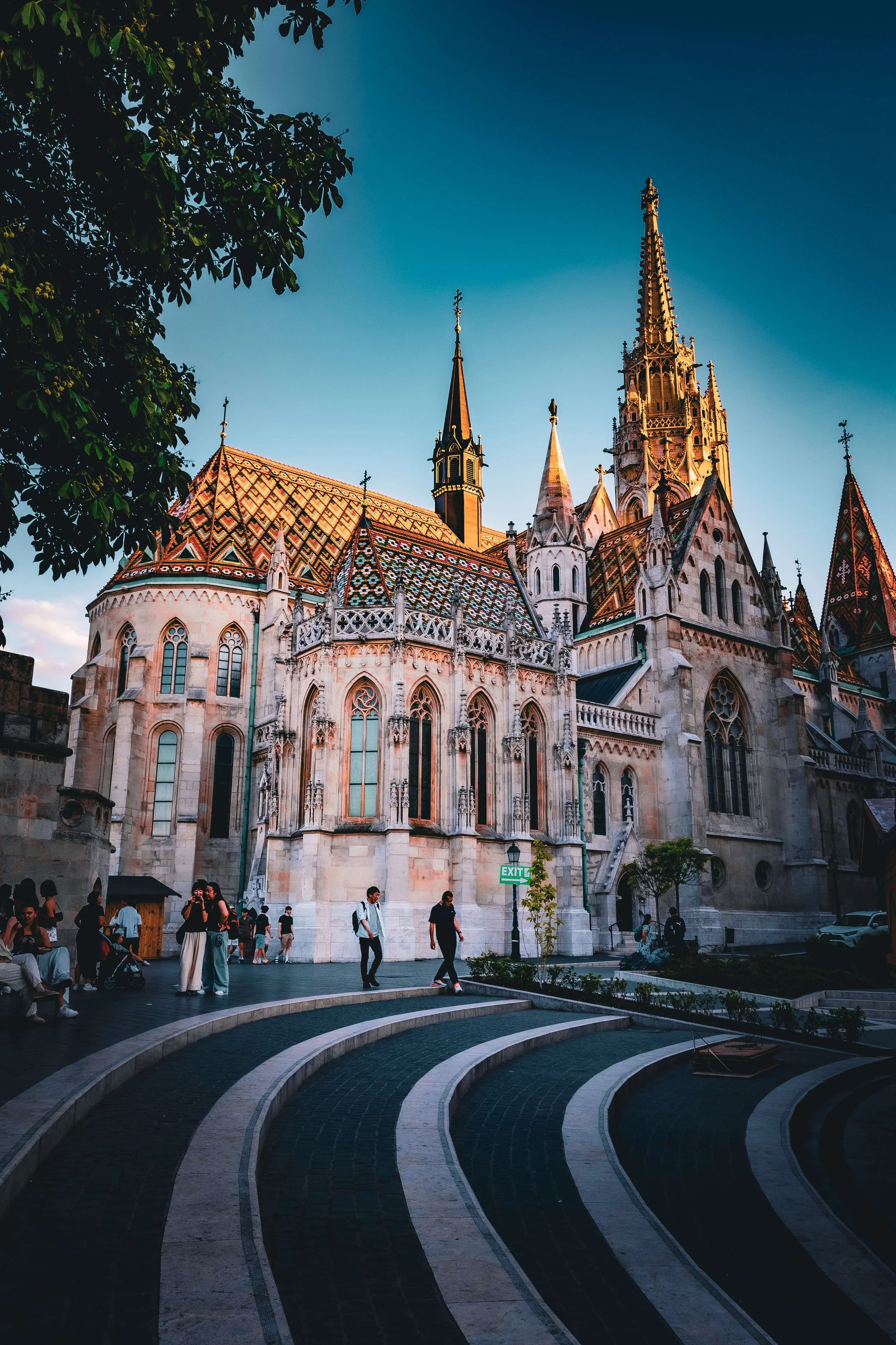 View of a Gothic-style church with colorful tiled roof, tall spires, and ornate architecture, sunlit against a clear blue sky, with people gathered in a town square in front.