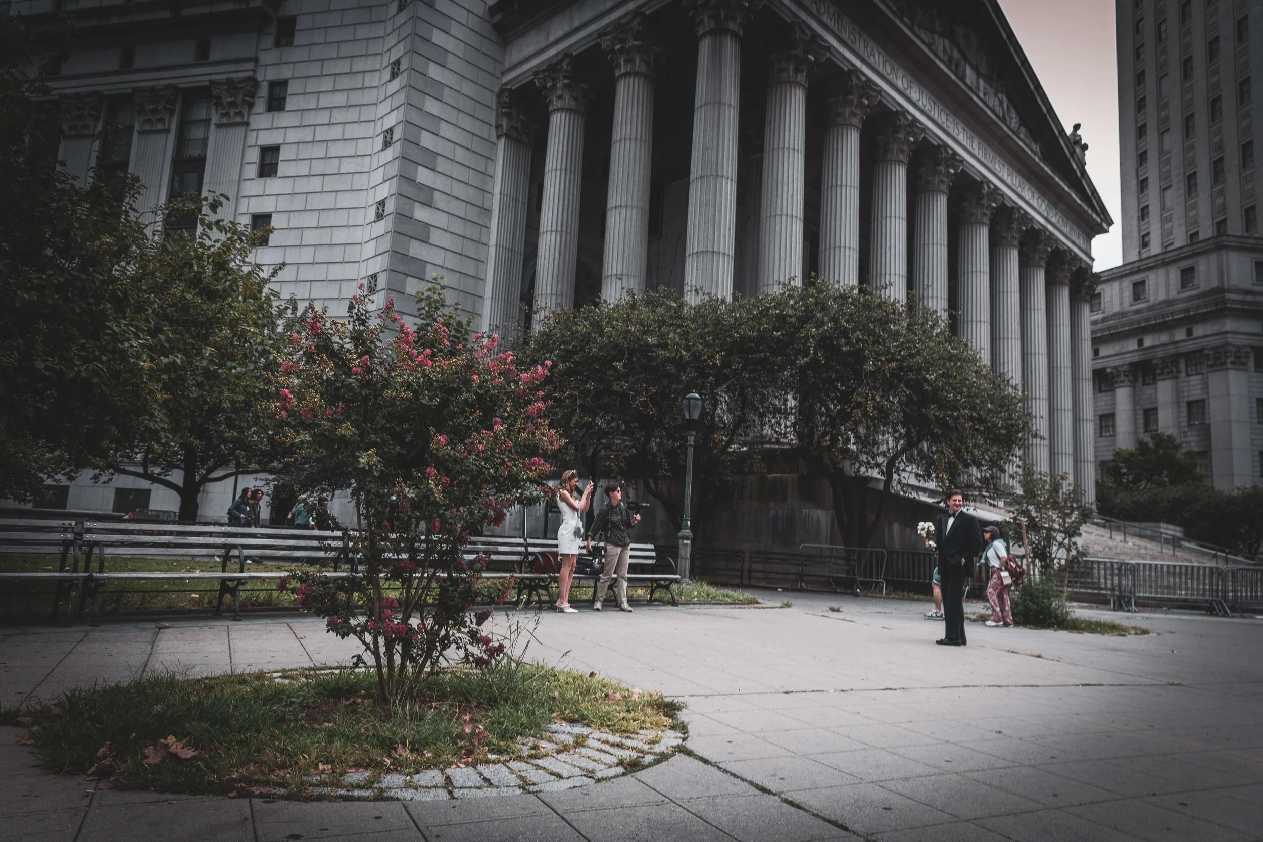 City street scene with a large classical courthouse featuring tall columns, people taking photos, and trees with pink flowers.