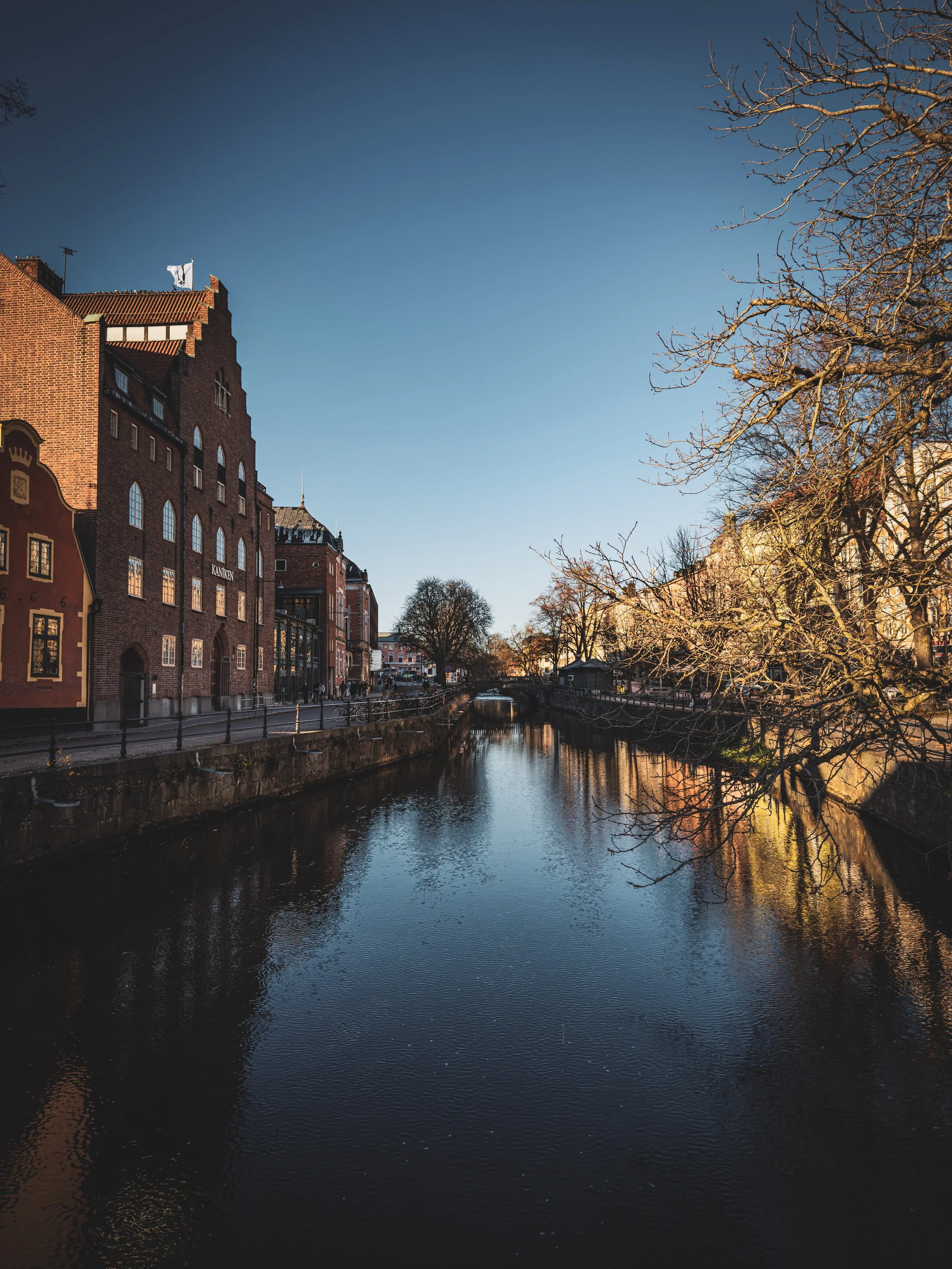 A calm canal lined with brick buildings on the left and leafless trees on the right under a clear blue sky.