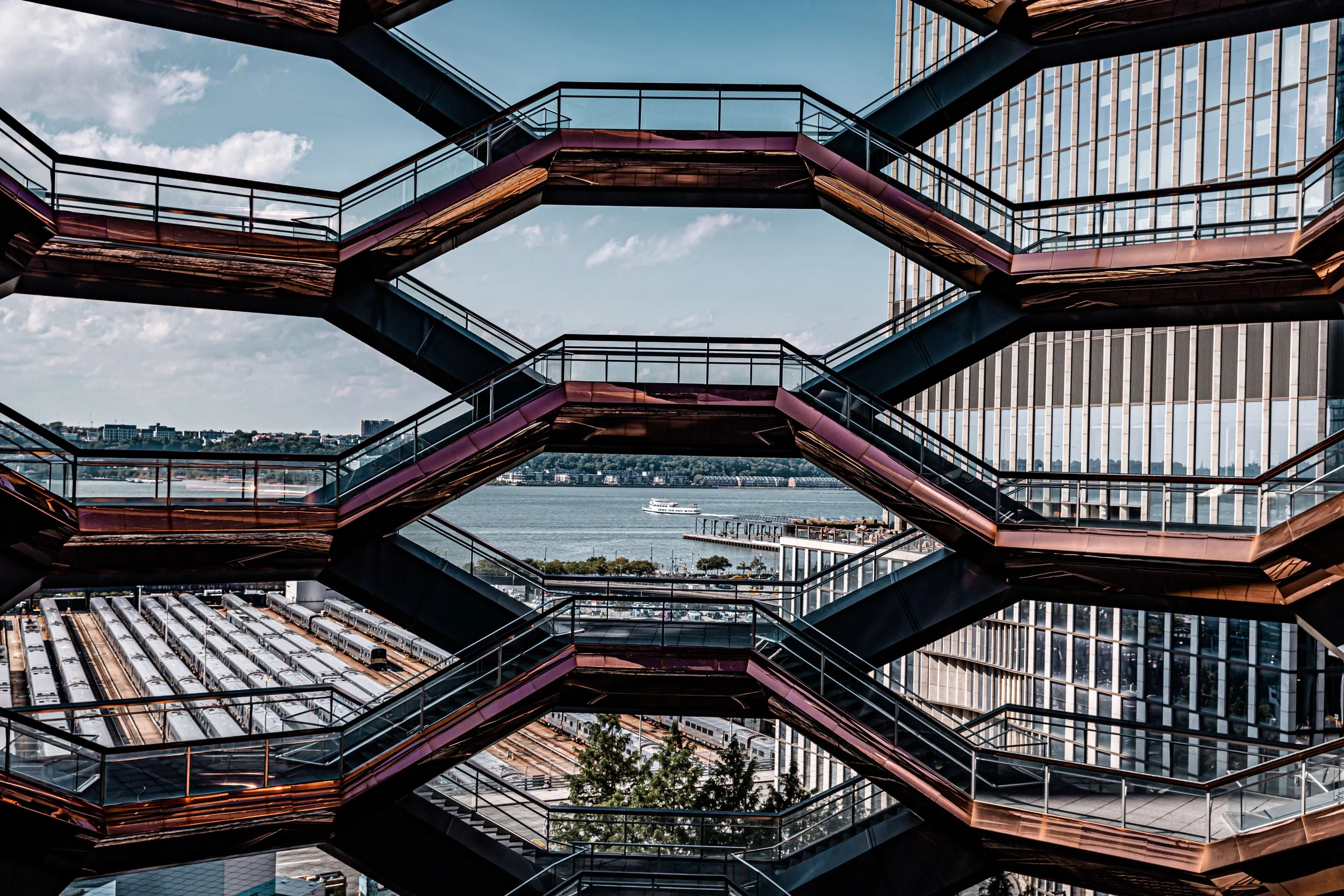 Exterior view of a modern building with multiple interconnected glass and steel staircases leading upwards, overlooking a cityscape with water, boats, and a bridge.