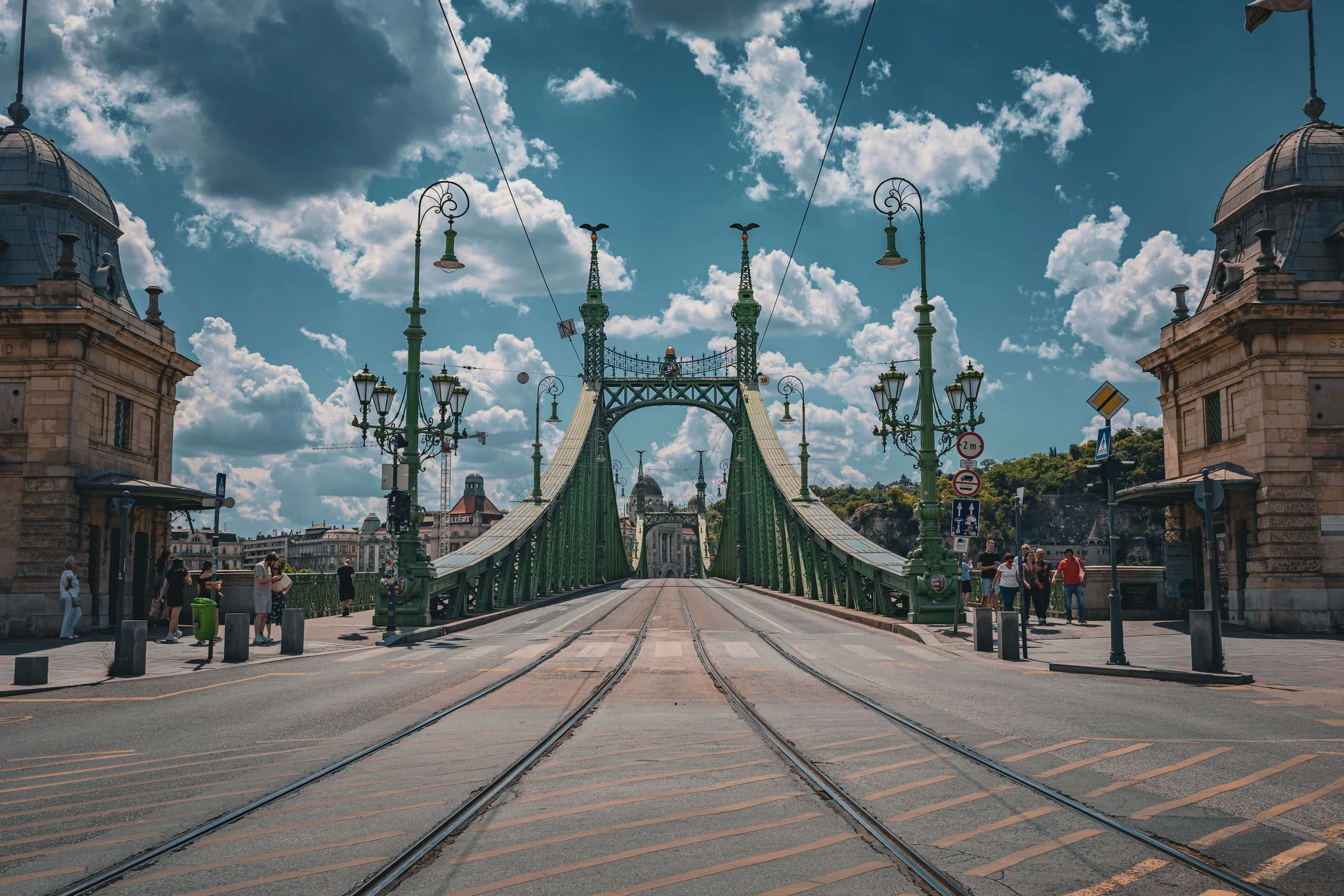 View of a bridge with tram tracks, ornate street lamps, and historic buildings under a partly cloudy sky in a cityscape.