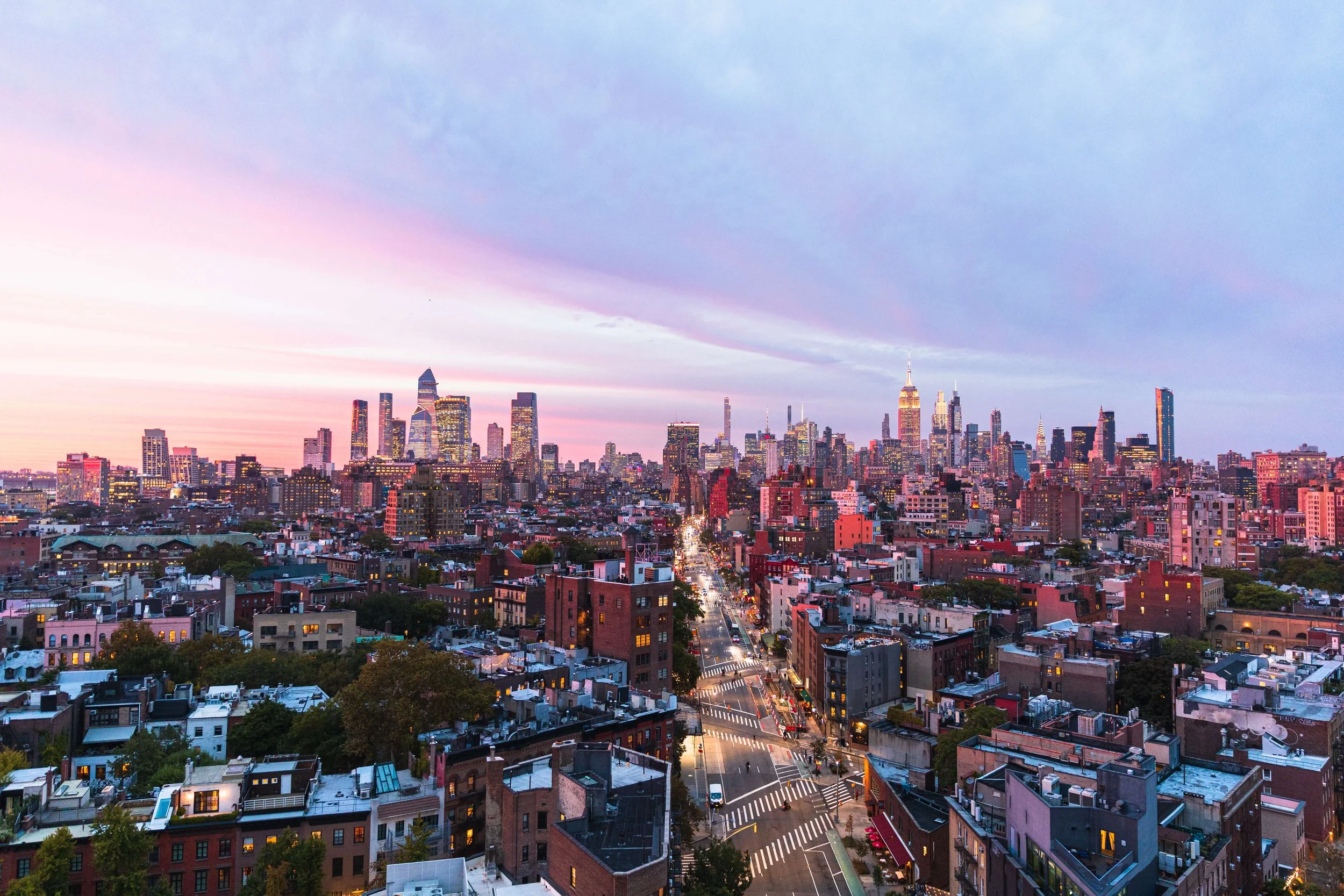 Sunset view of the New York City skyline with tall skyscrapers, including the Empire State Building, under a sky with pink and blue streaks, and busy streets with cars below.