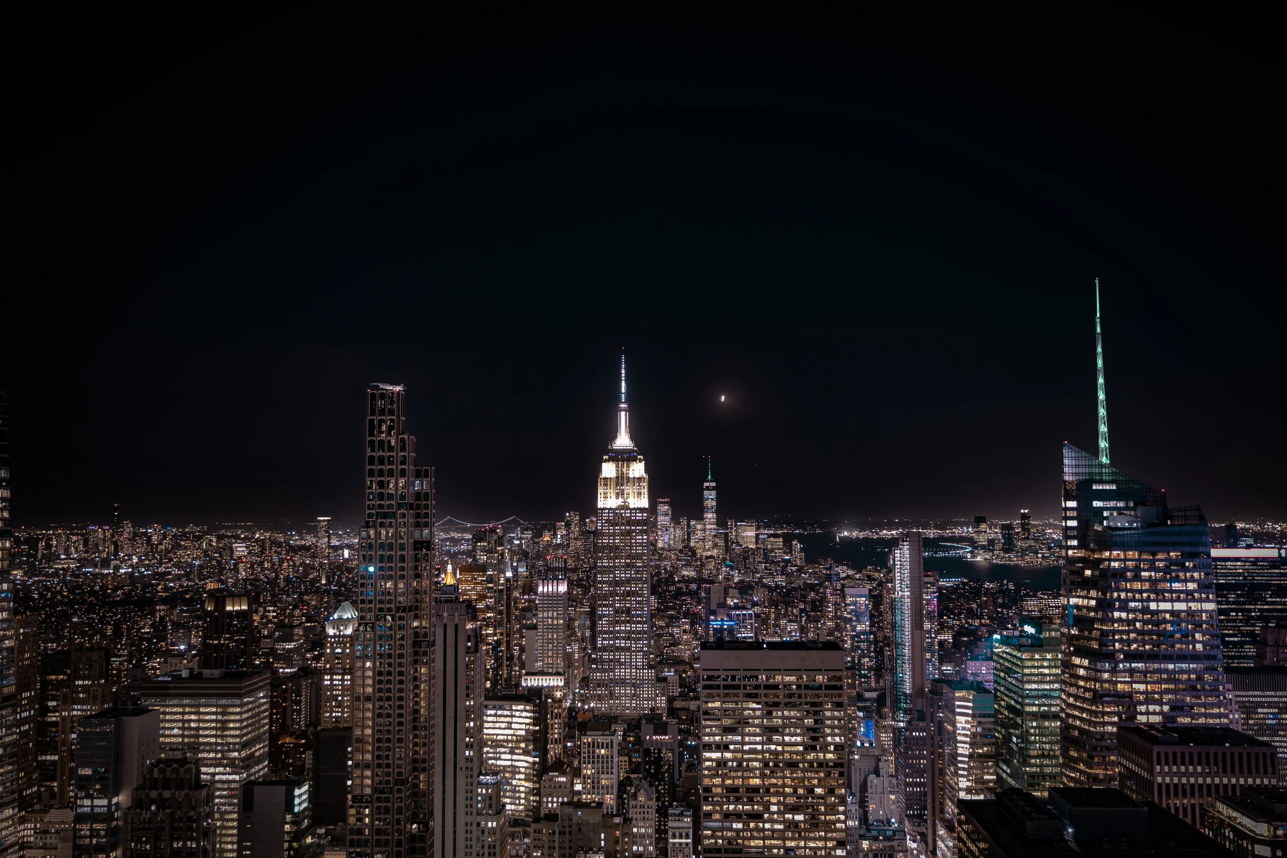 Nighttime cityscape of New York City with illuminated skyscrapers, including the Empire State Building, under a dark sky with a visible moon.